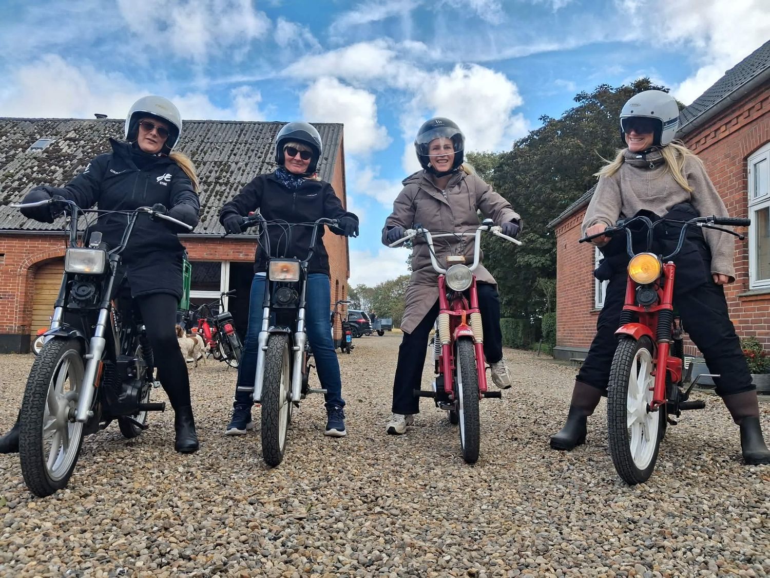 Four people wearing helmets pose on vintage mopeds on a gravel path beside a brick building on a sunny day.