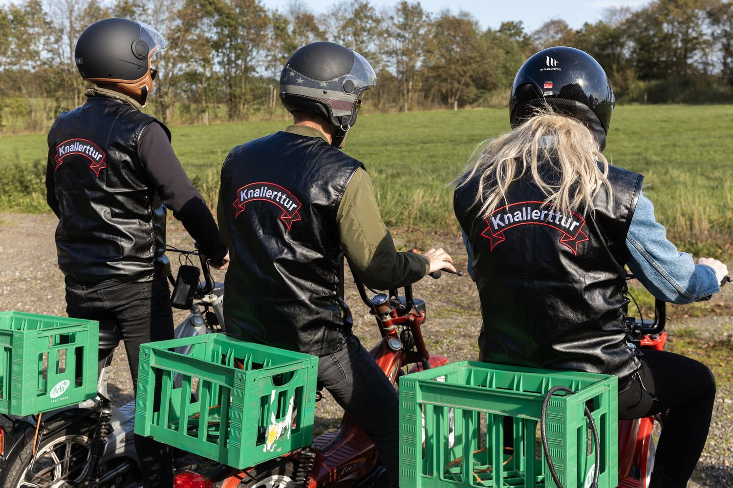 Three motorcyclists wearing Knallerttur vests ride bikes with green crates on a rural road.