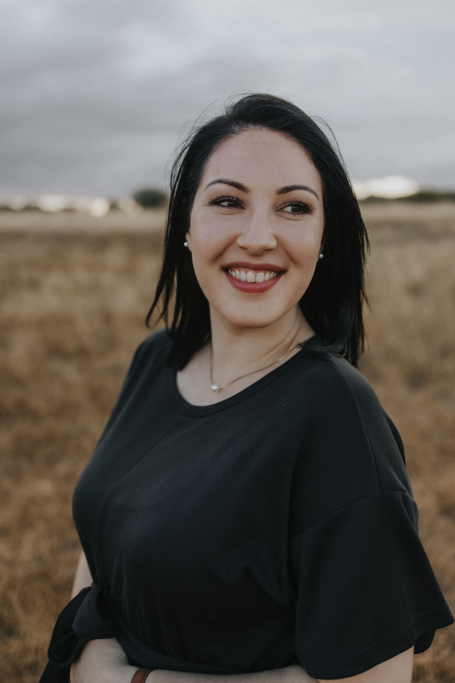 Outdoor portrait of woman in black top smiling against rural landscape.
