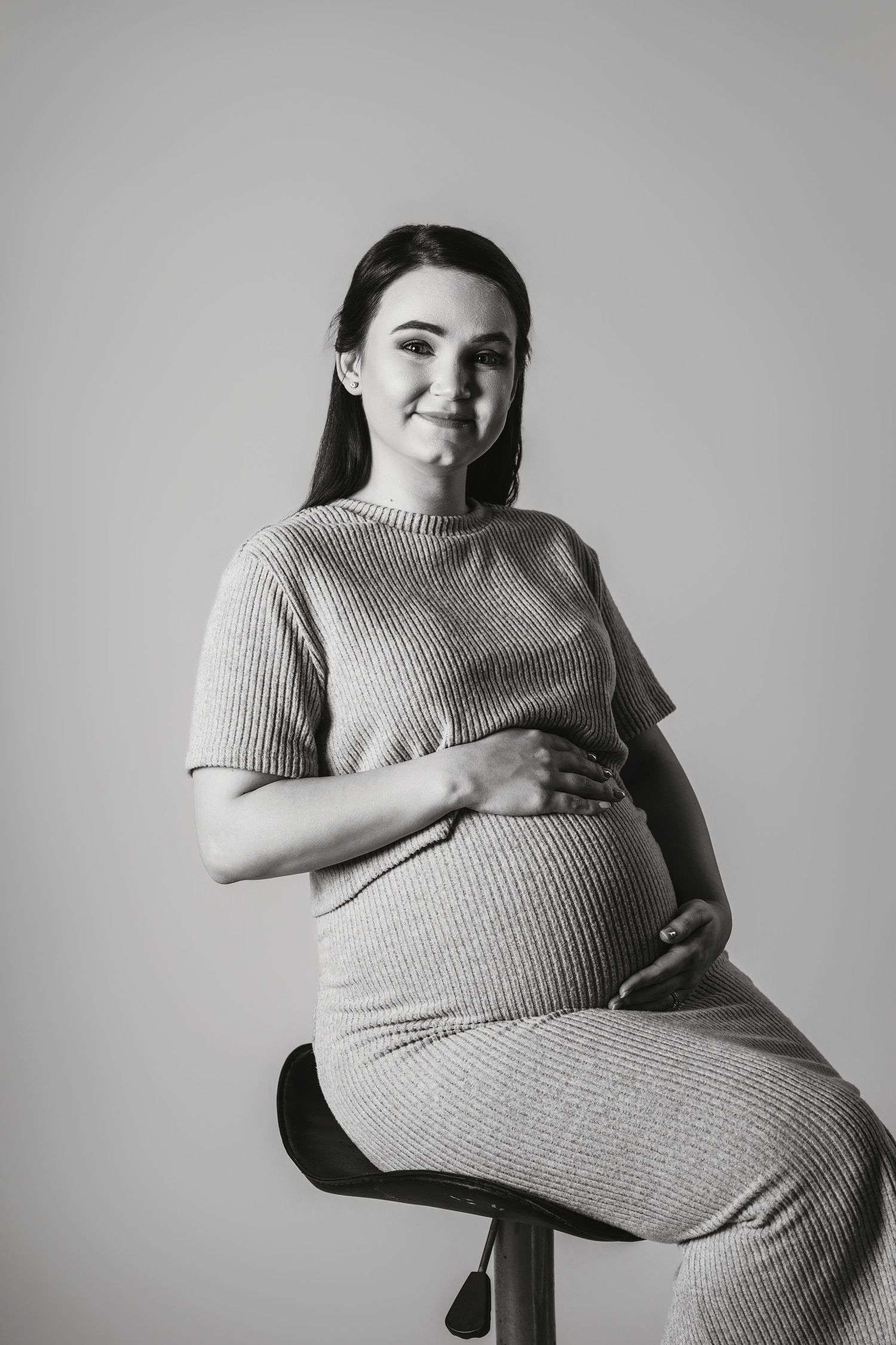 Black and white artistic maternity photo series showing a pregnant figure in a ribbed sweater reclining on a chair.