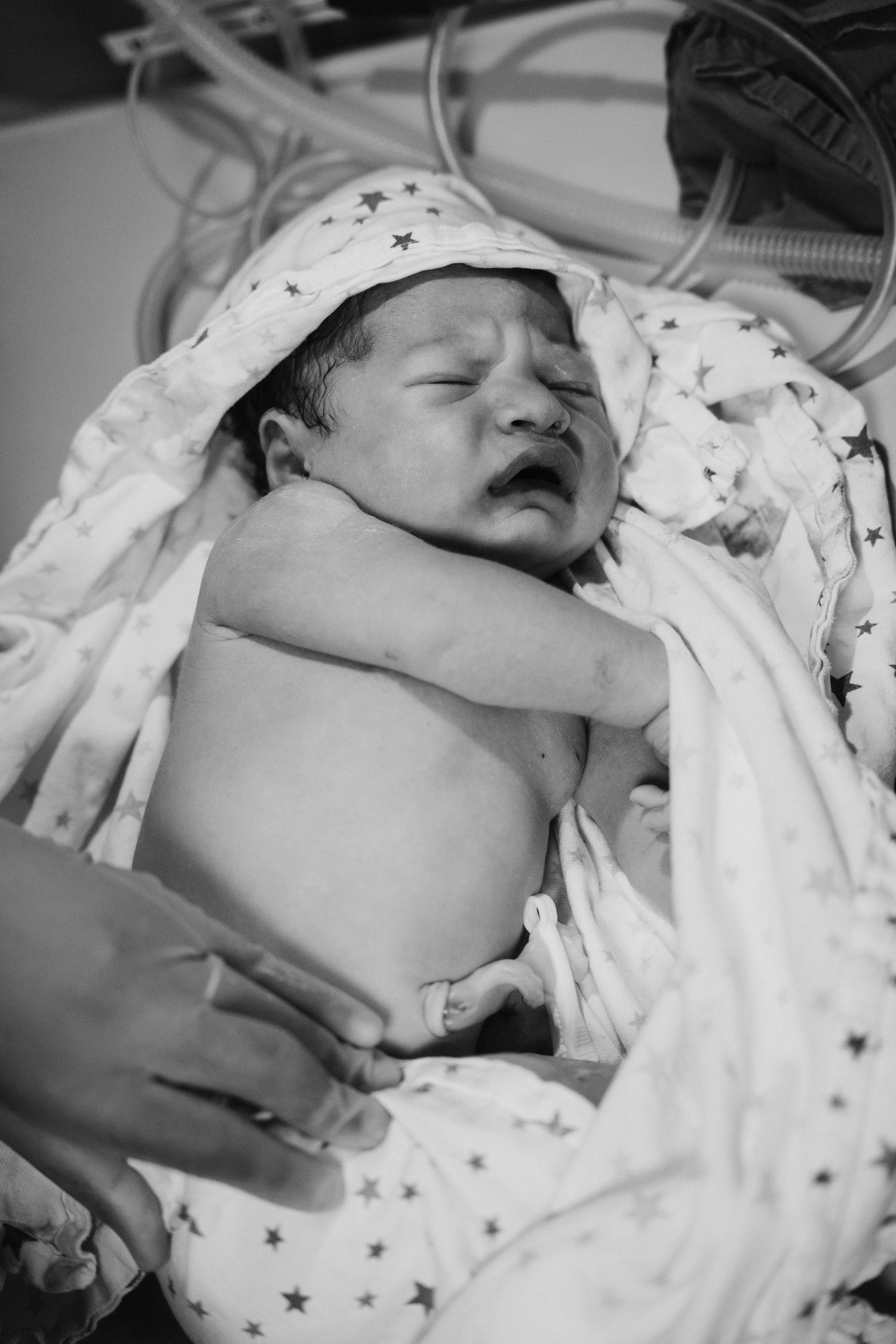 Black and white photo of a newborn baby swaddled in star-patterned blanket resting in hospital bed.
