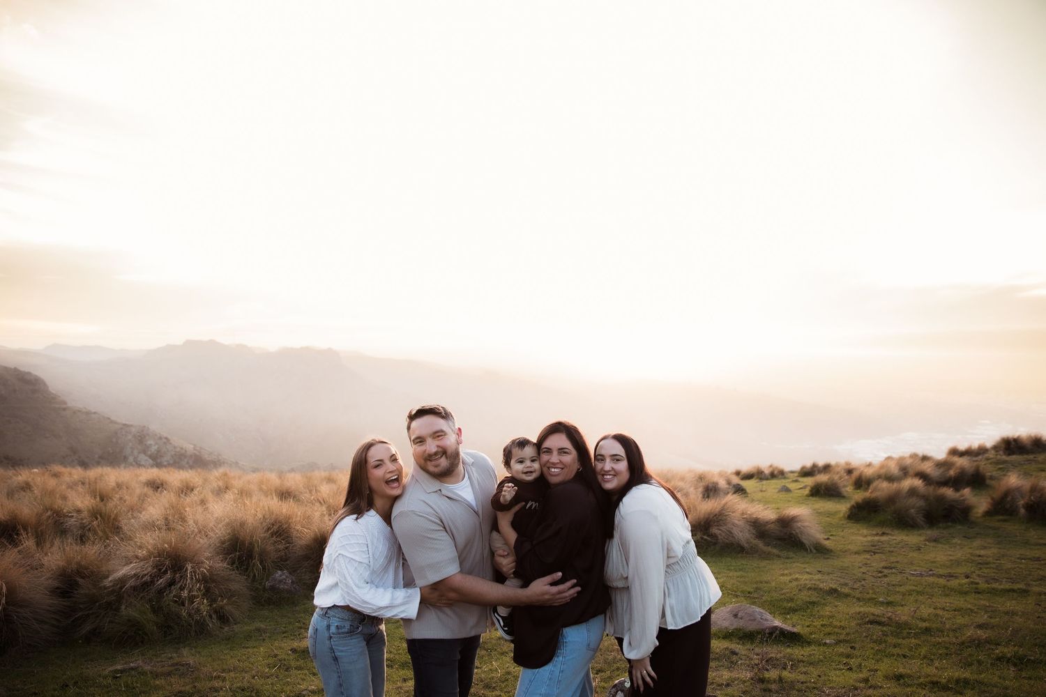 Family of five smiling together on a scenic hilltop at golden hour with mountain views in the background.