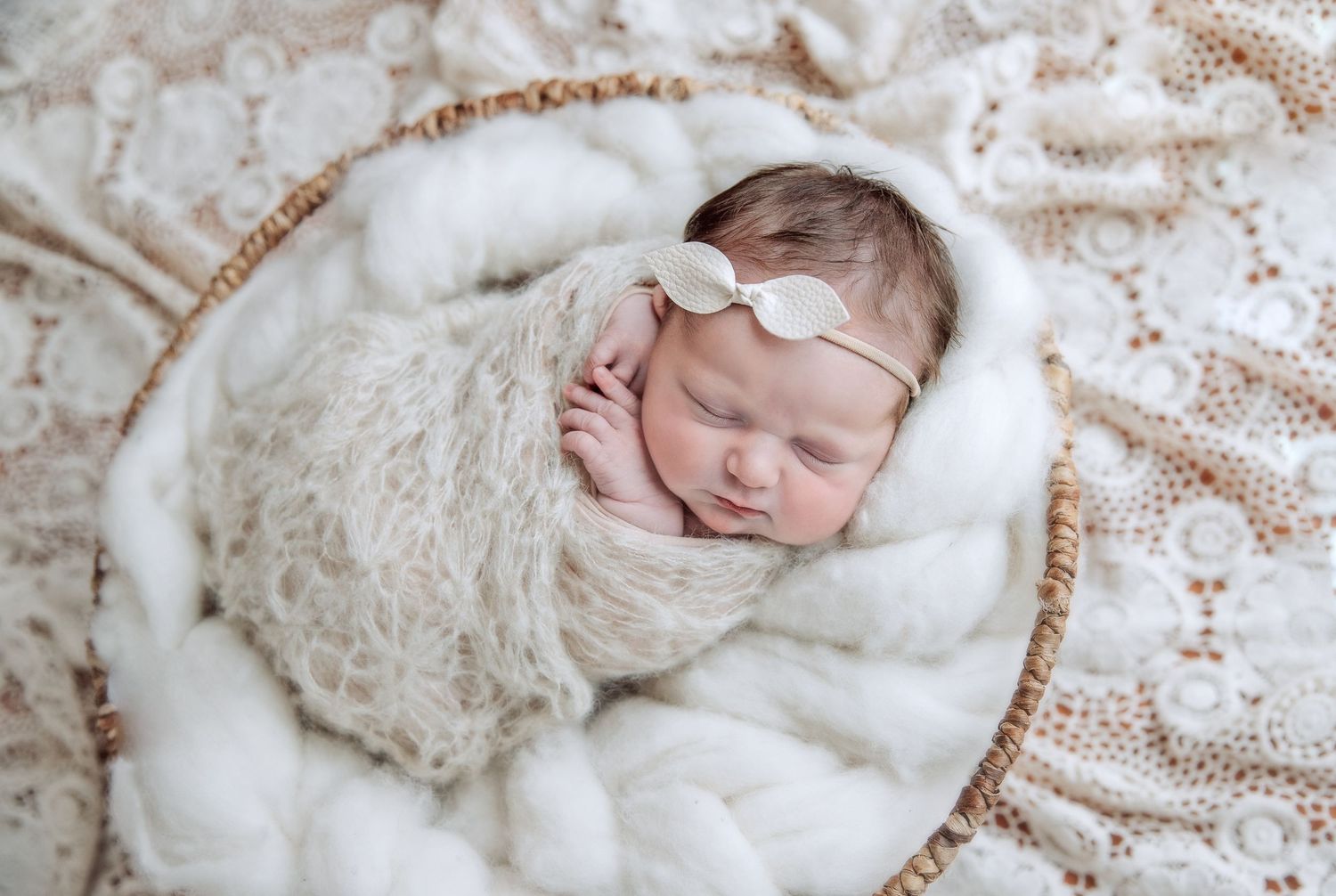 Sleeping newborn wrapped in soft white lace and fur with delicate bow headband against floral backdrop.