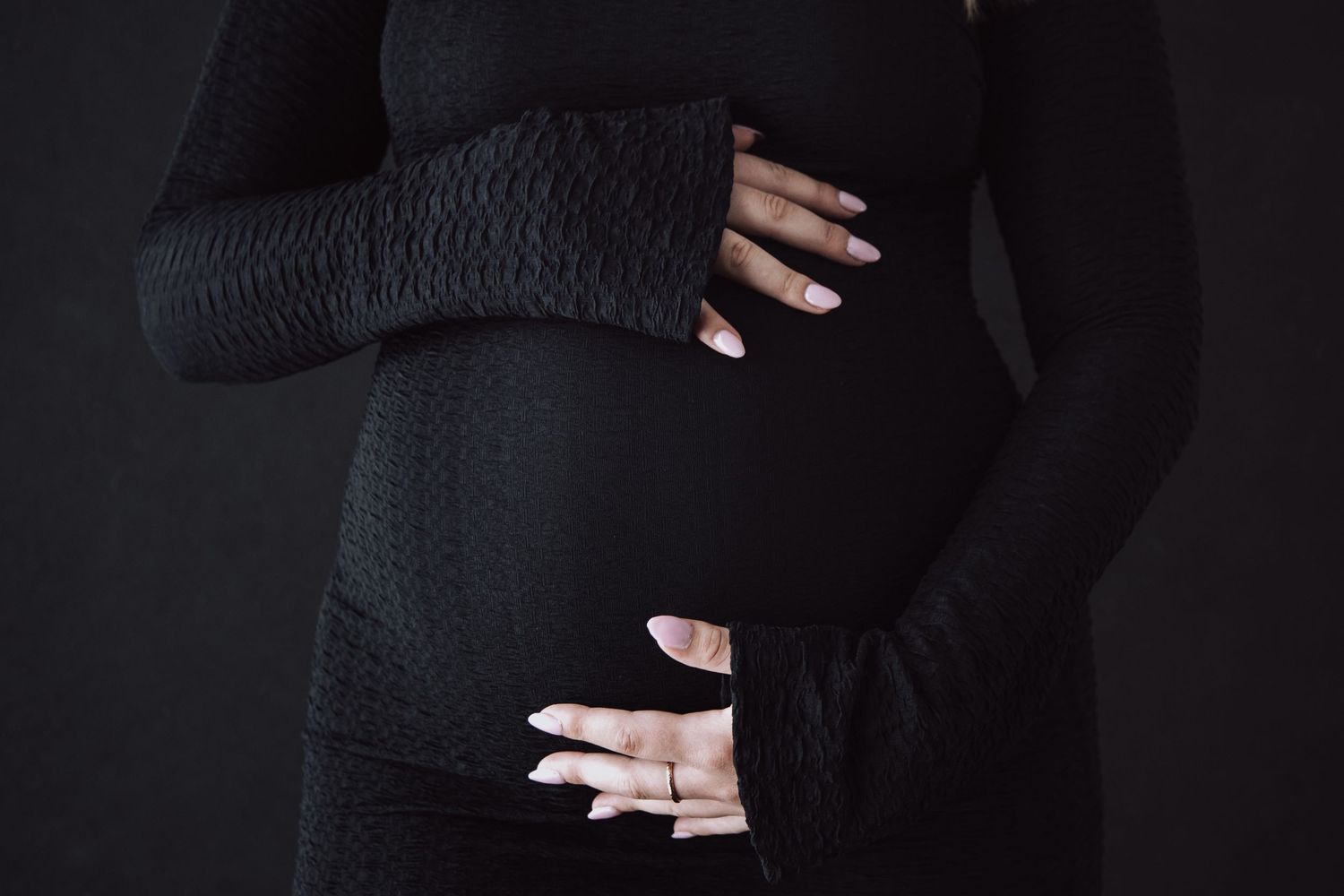 Hands with long white nails rest on a pregnant belly covered in a black knit sweater against a dark background.