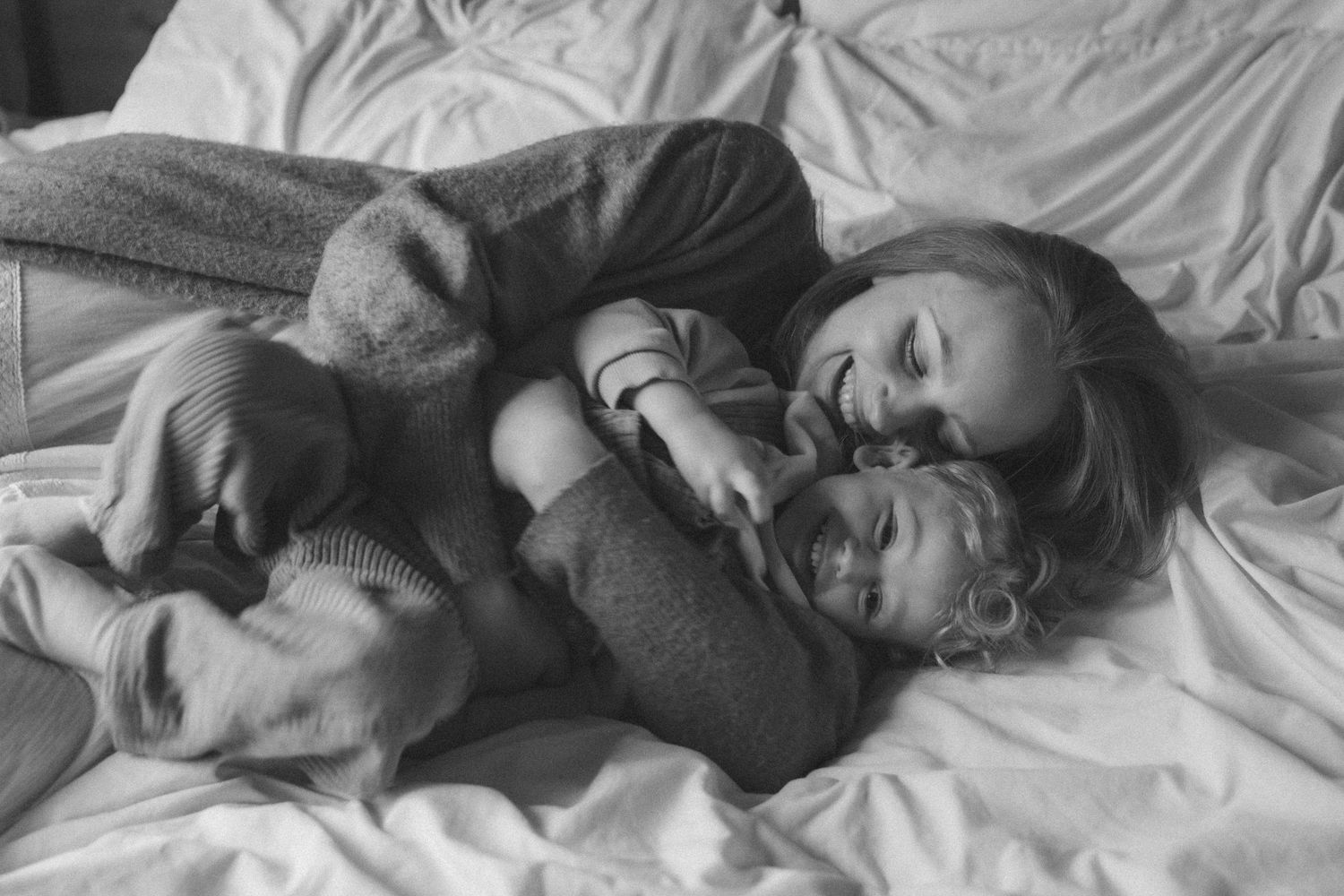 Black and white photo of a mother snuggling with her two young children on a white bed.