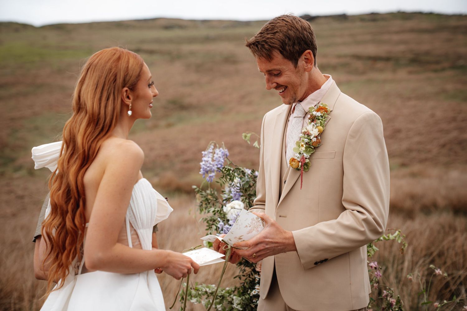 Bride and groom exchange vows outdoors on a moorland, groom in beige suit holding wildflowers.