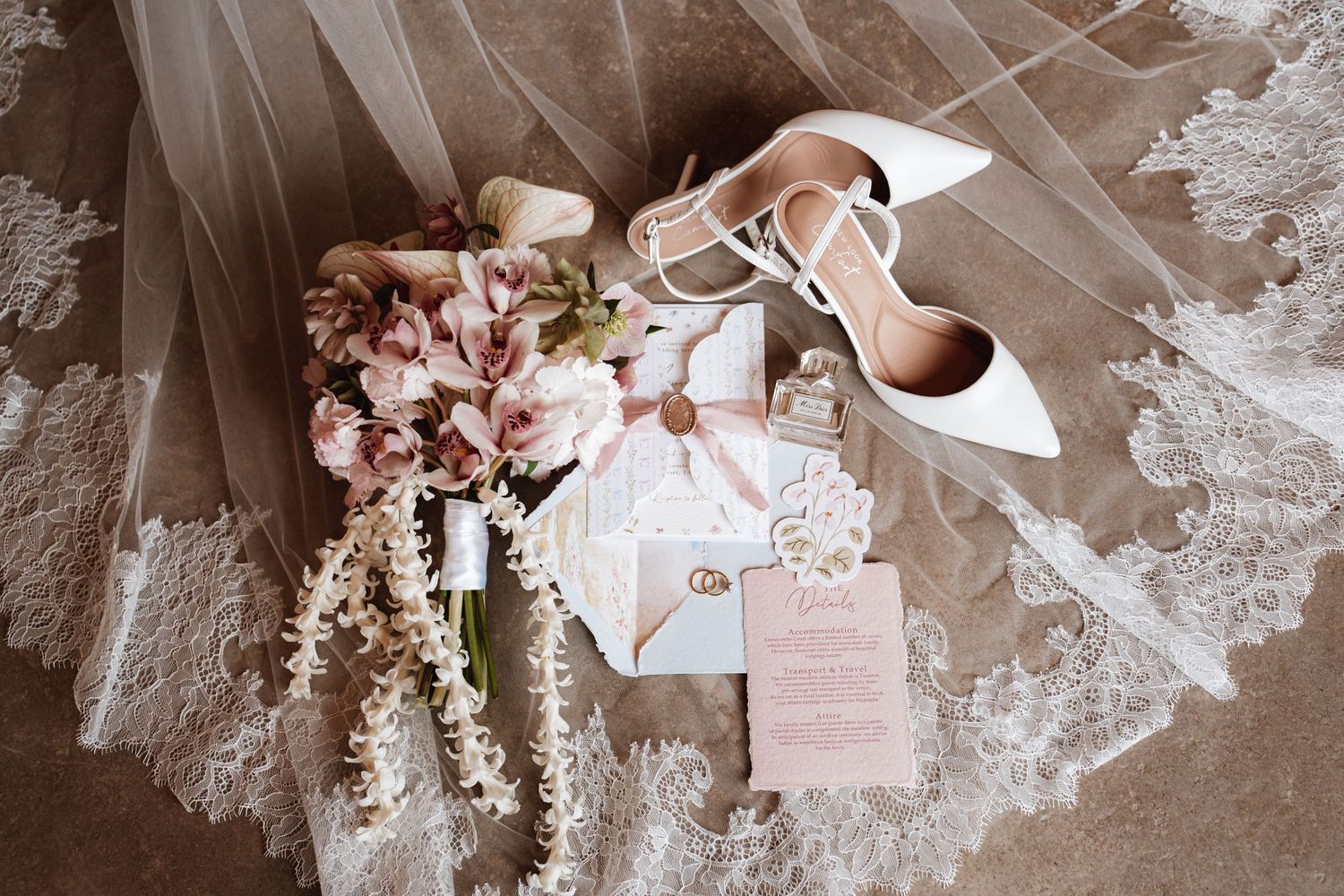 Flat lay of wedding details including bouquet, heels, rings, and invitation suite on lace veil.