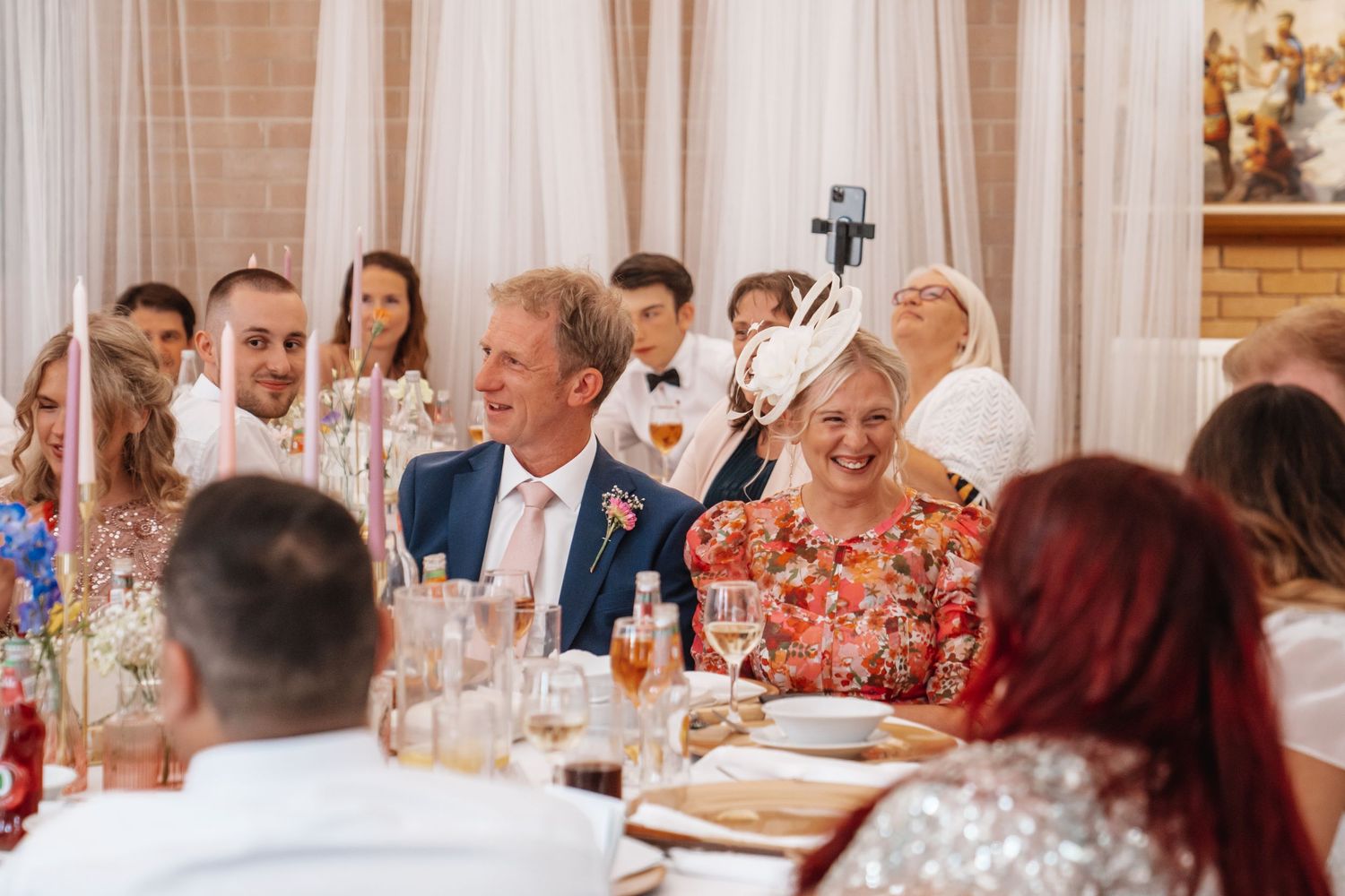 Wedding guests laugh and take selfies at elegantly decorated reception venue with floral centerpieces.