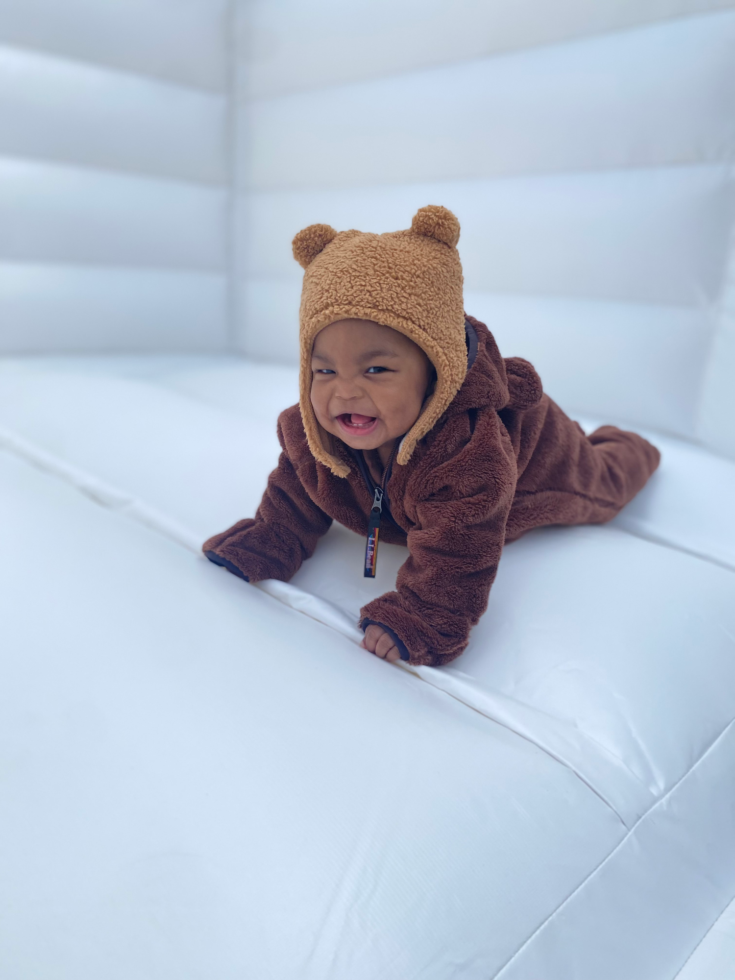 A happy baby wearing a brown bear costume lying on a white inflatable surface.