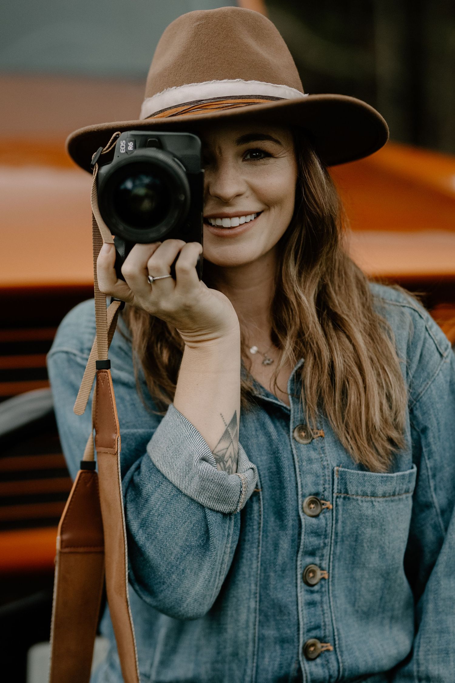 Photographer wearing denim jacket and brown fedora holds camera while smiling during outdoor portrait session.