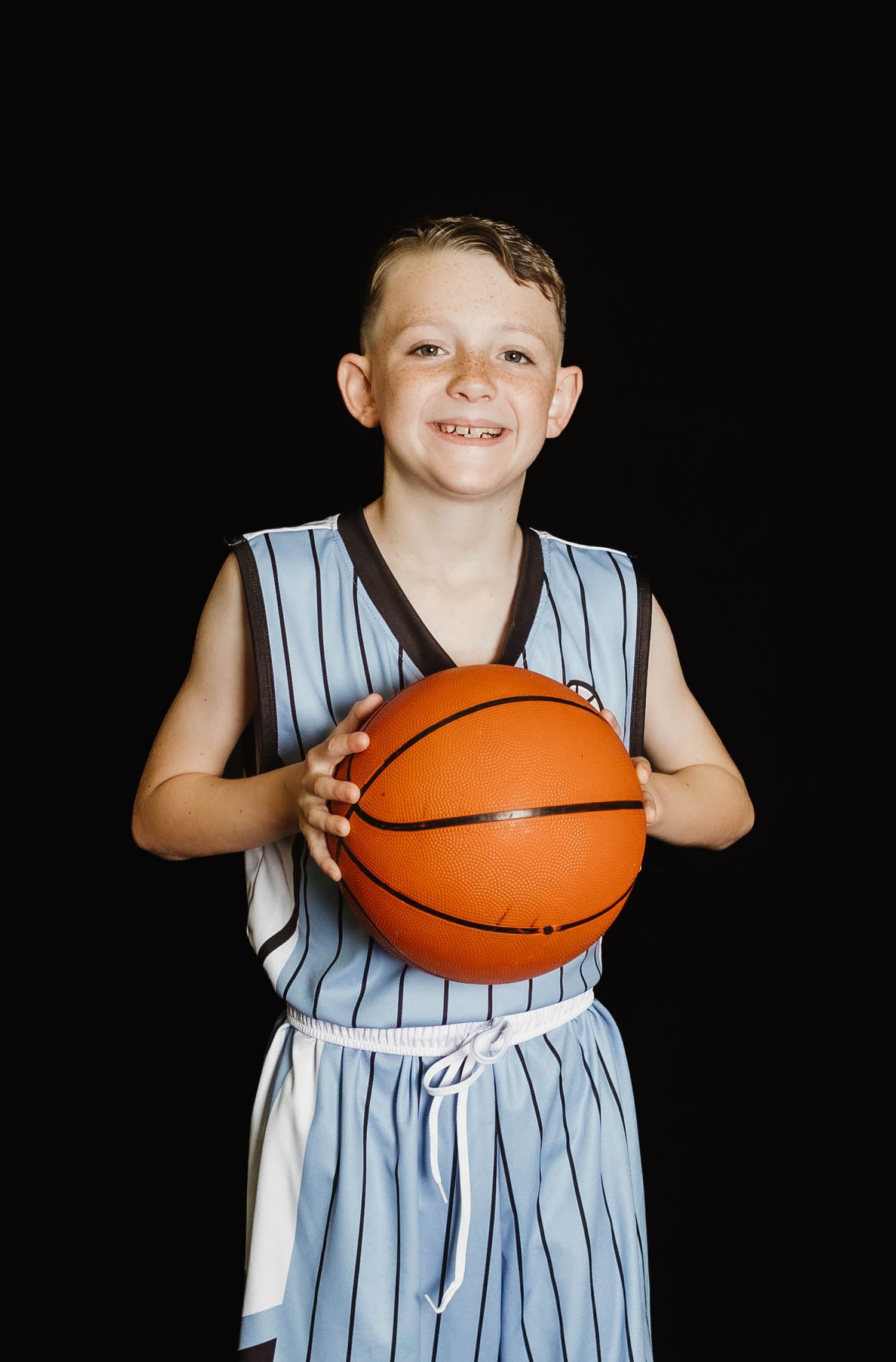 Young basketball player in light blue and white striped uniform holds orange basketball against dark background.