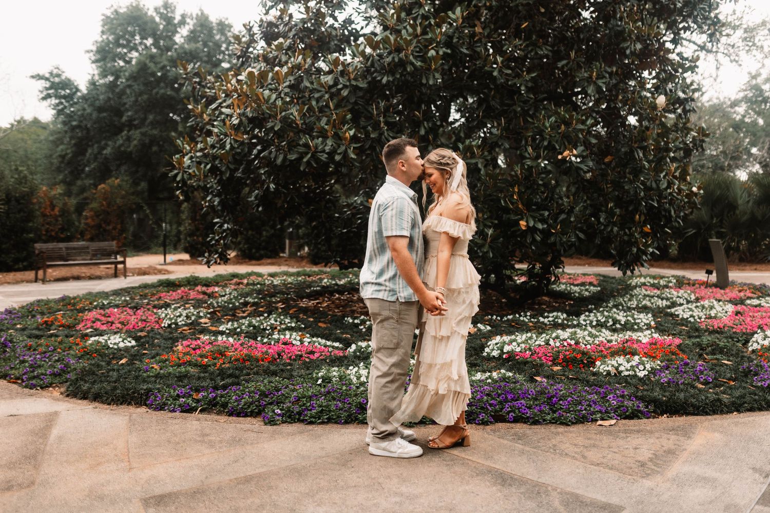 A couple shares an intimate moment in a beautiful garden surrounded by colorful flower beds and trees.