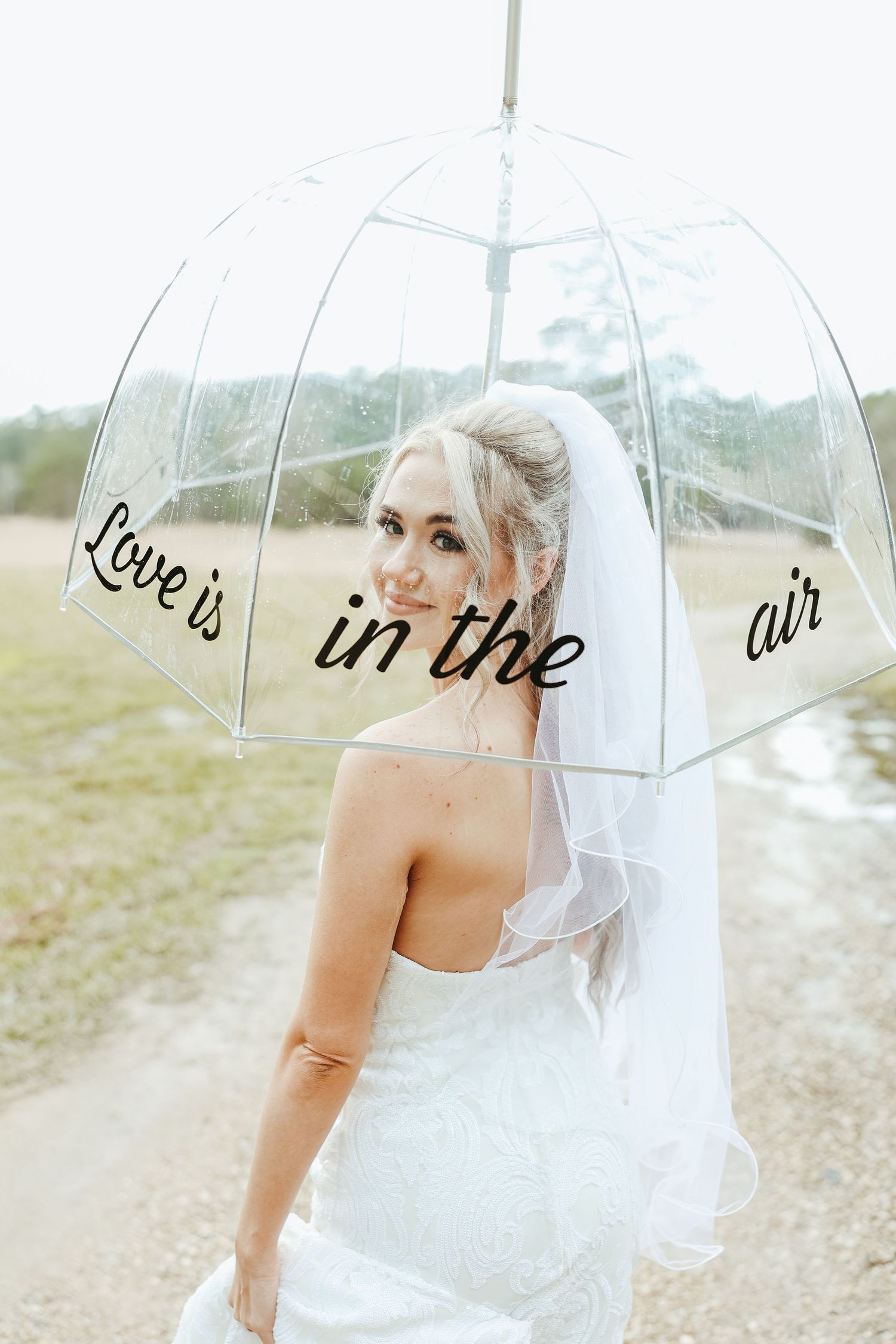 Clear umbrella with 'love is in the air' text held by bride in white wedding dress with flowing veil outdoors.