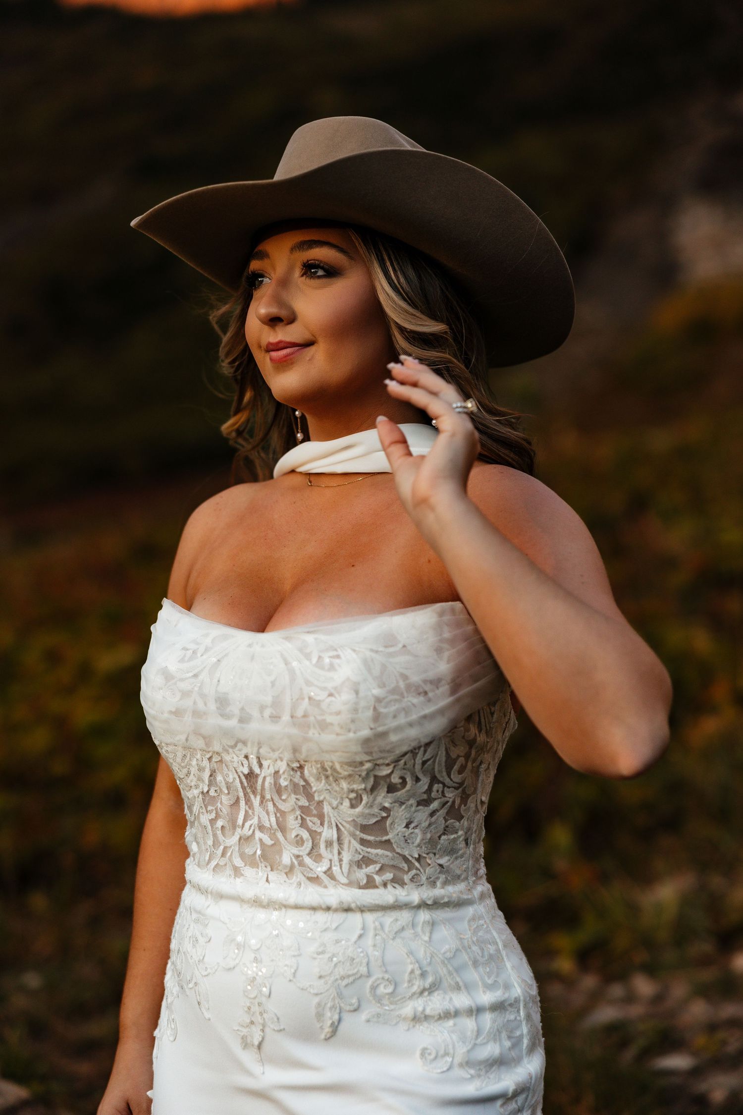 A bride in a white lace wedding dress and wide-brimmed hat poses outdoors during a sunset photoshoot.