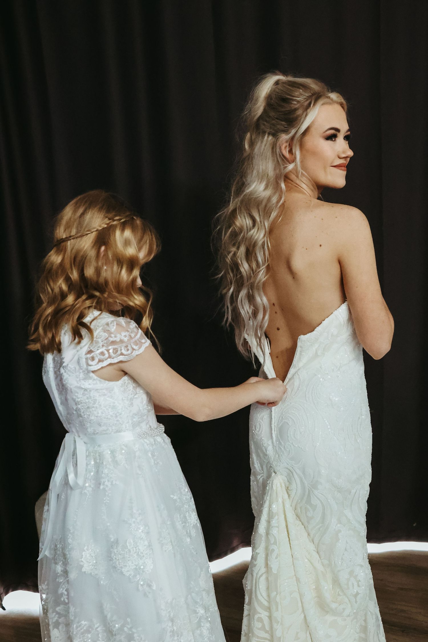 Two people in white lace dresses with flowing hair stand together in an intimate wedding preparation moment.