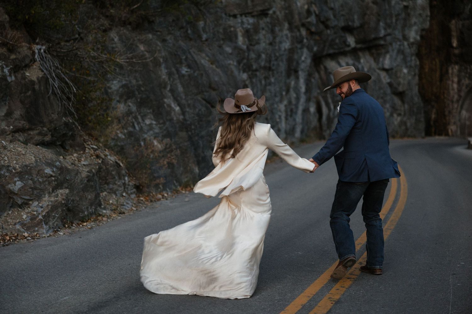 Couple dances on mountain road with white dress flowing as they spin together against rocky cliffs.