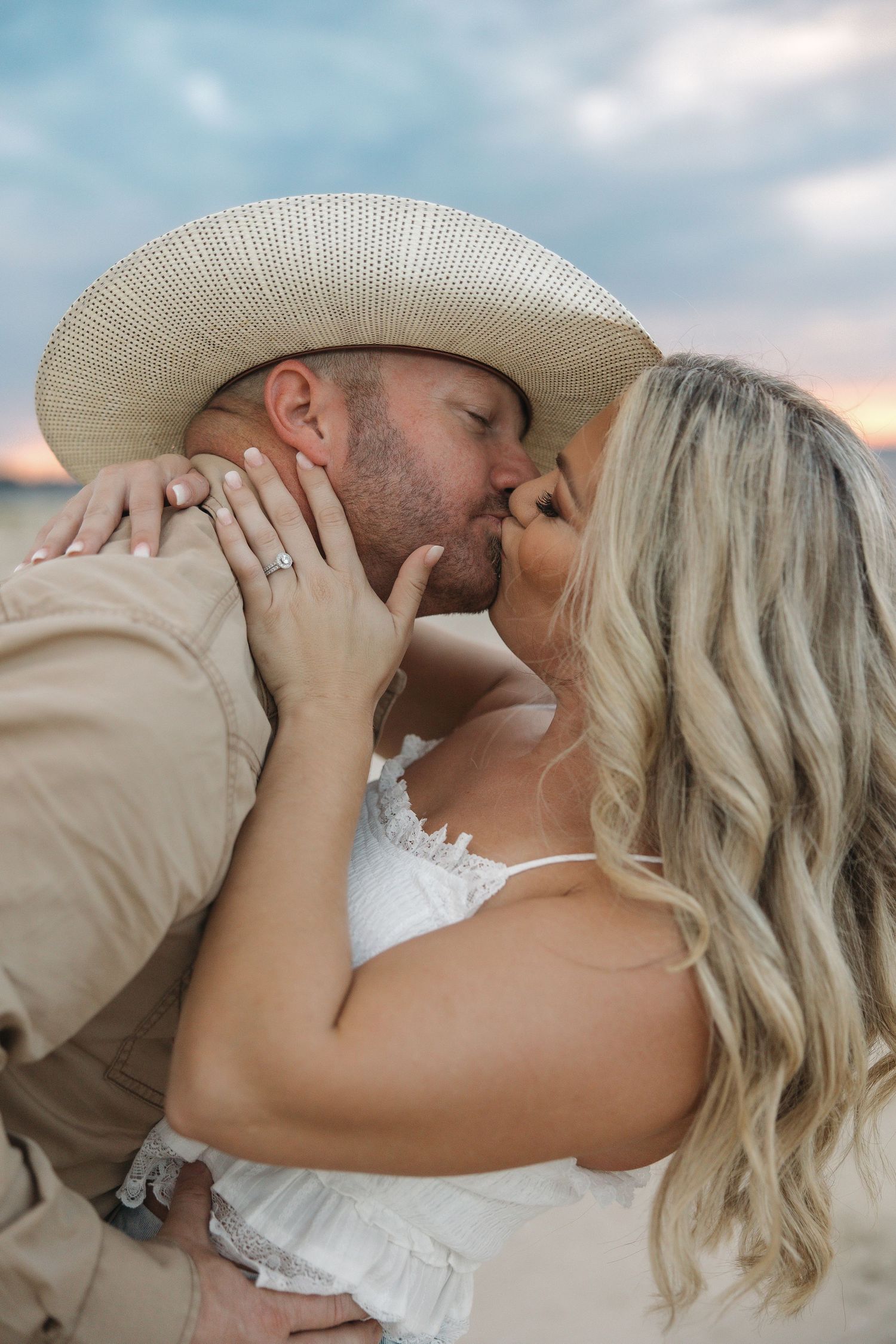 Romantic couple in western attire sharing intimate kisses against a cloudy sky backdrop.