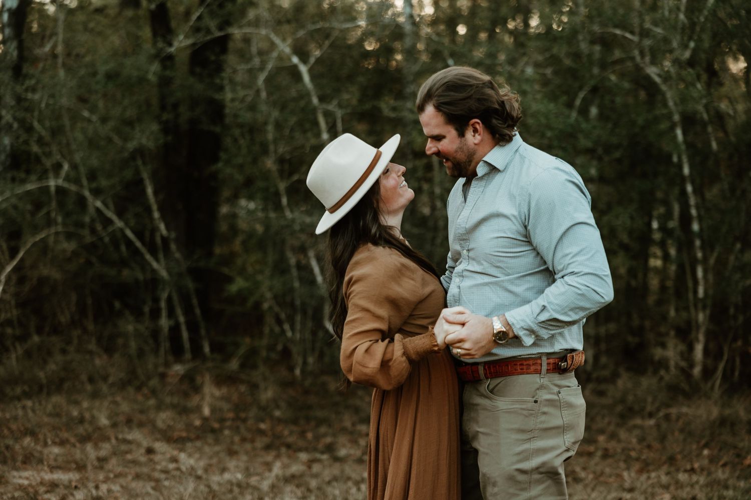 A couple shares an intimate moment in a wooded area, with her in a brown coat and him wearing a light blue shirt.