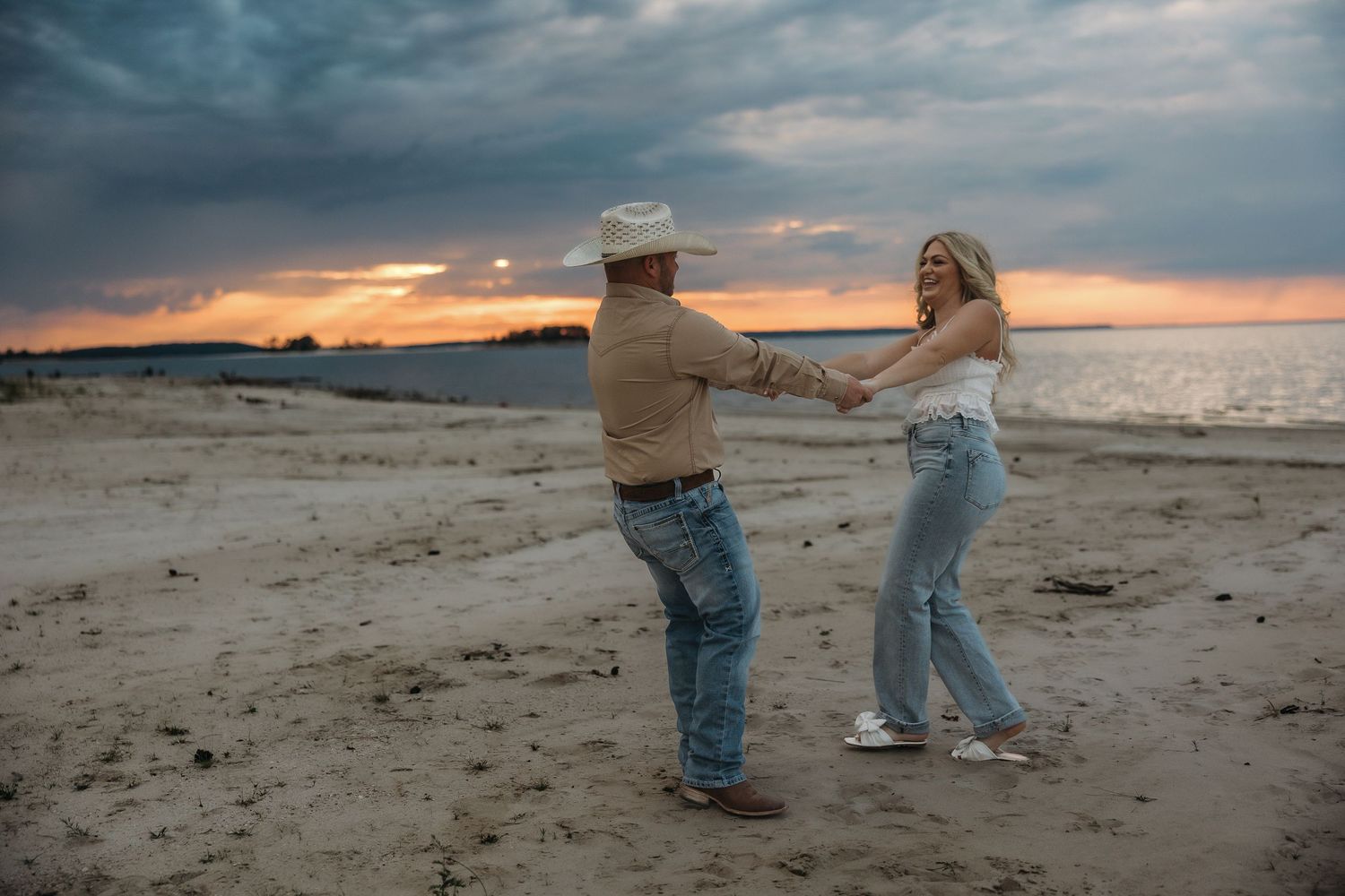 A couple dancing together on a sandy beach at sunset with dramatic cloudy skies overhead.