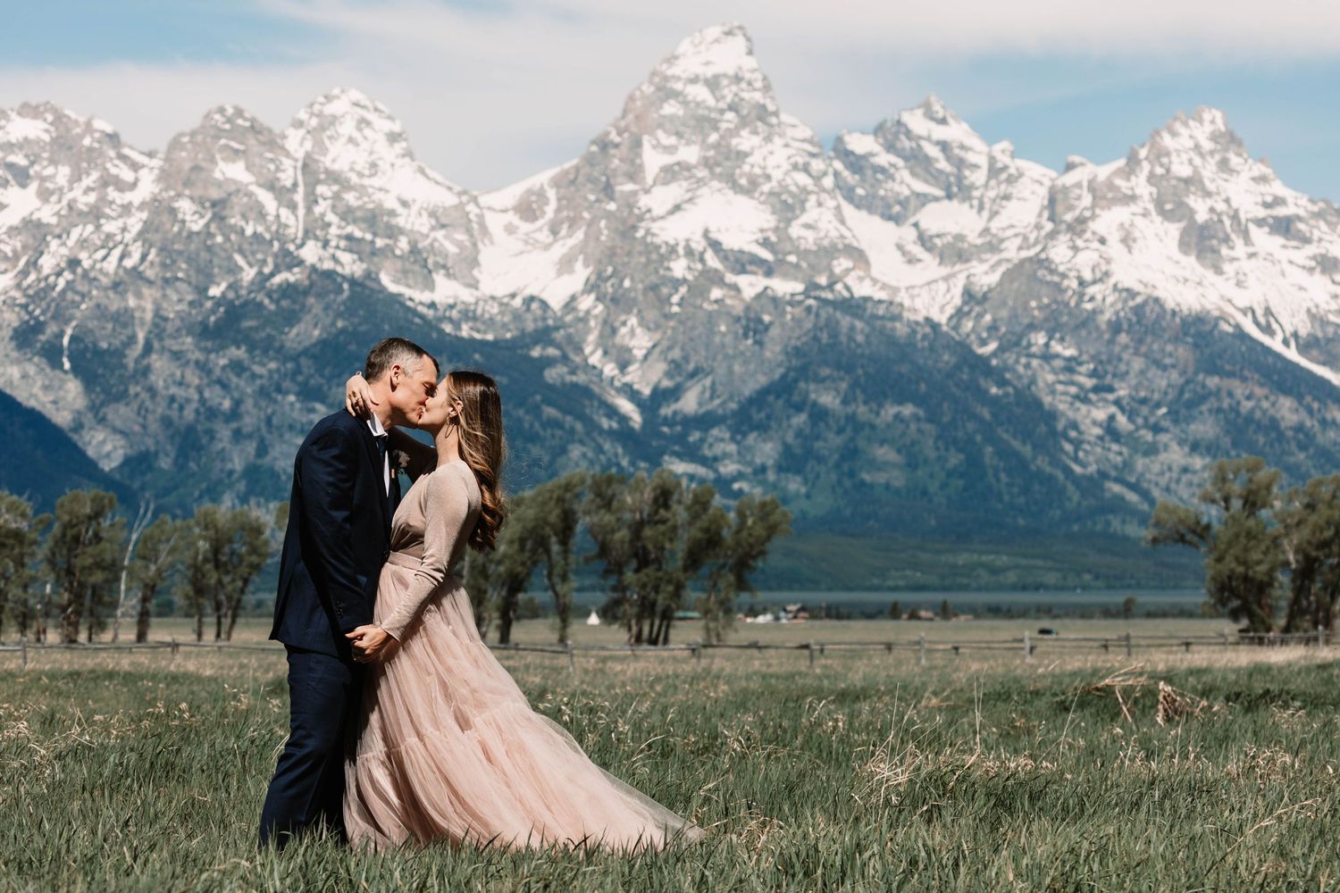 A couple embraces in a meadow with the majestic snow-capped Grand Teton mountains rising dramatically in the background.