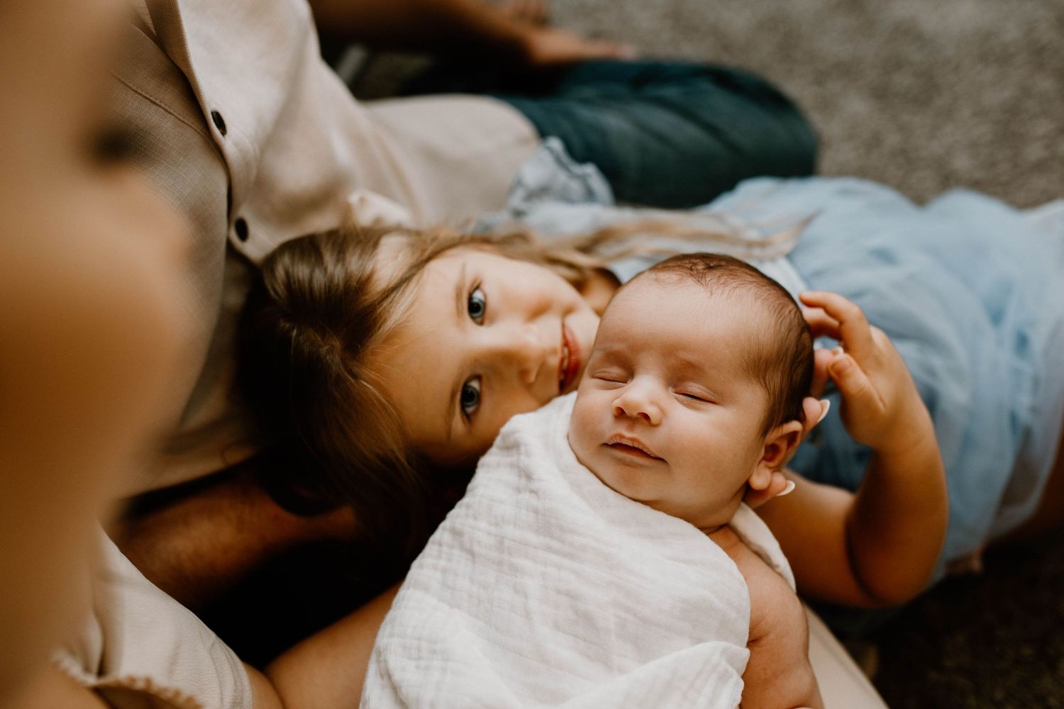 Two children lying down together on the floor, with the older child gently touching the baby's head.