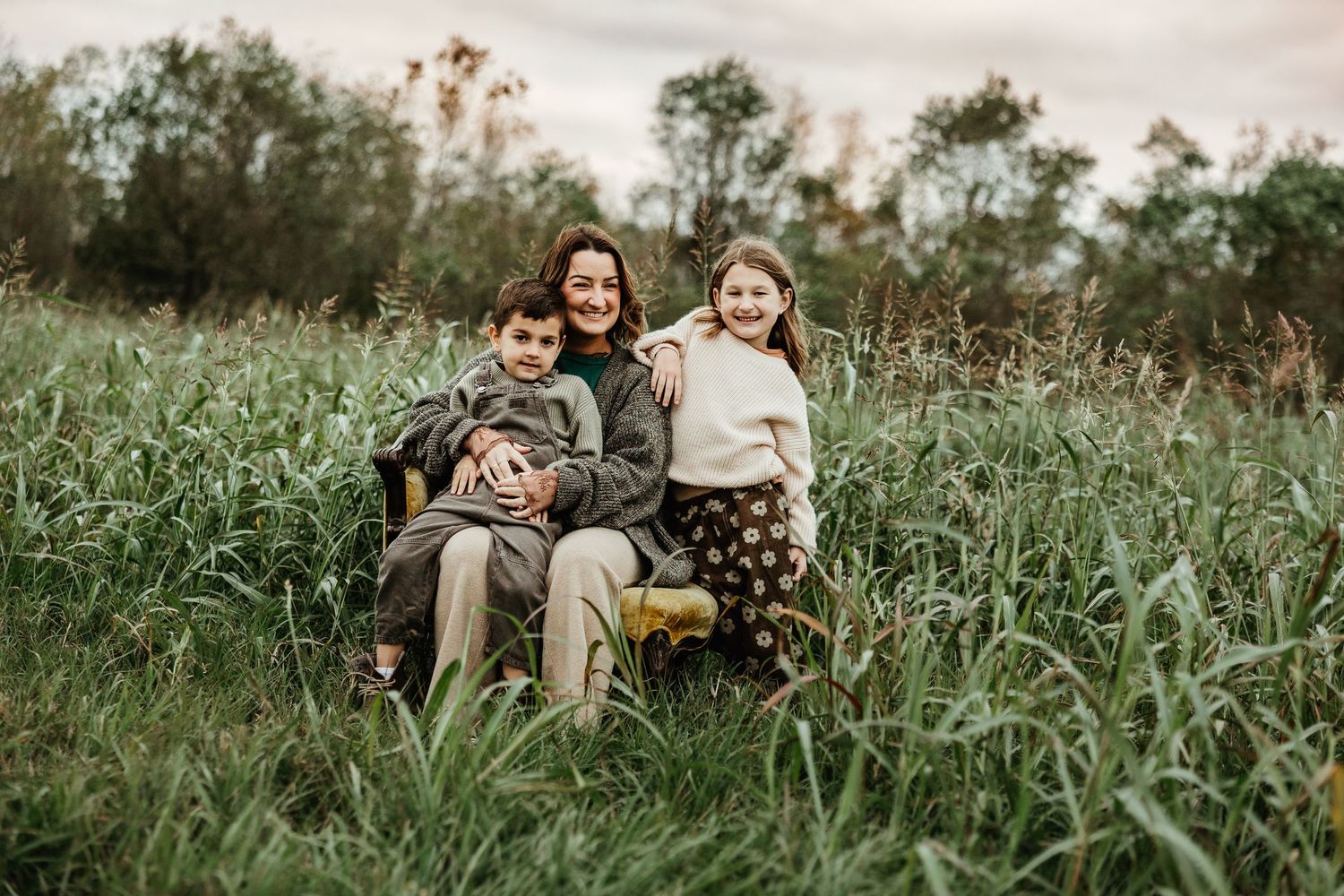 Three siblings sit together in tall grass during a candid outdoor photo session at golden hour.