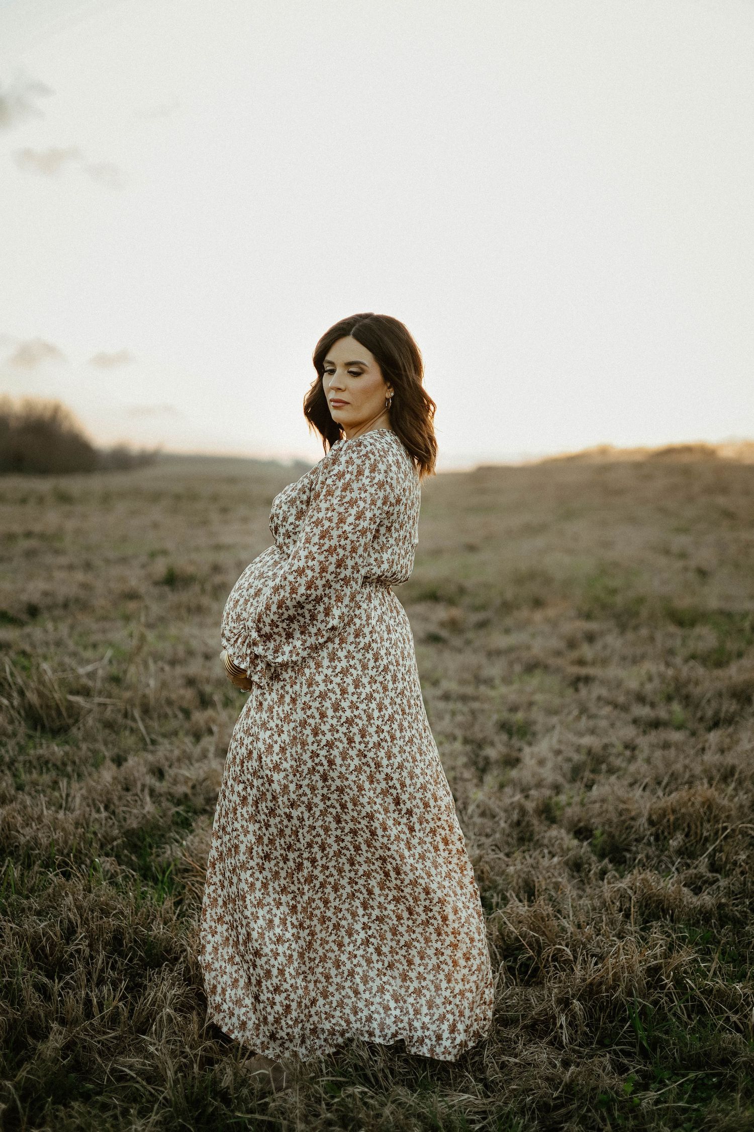 A pregnant woman in a flowing floral dress stands on a grassy hill at sunset.