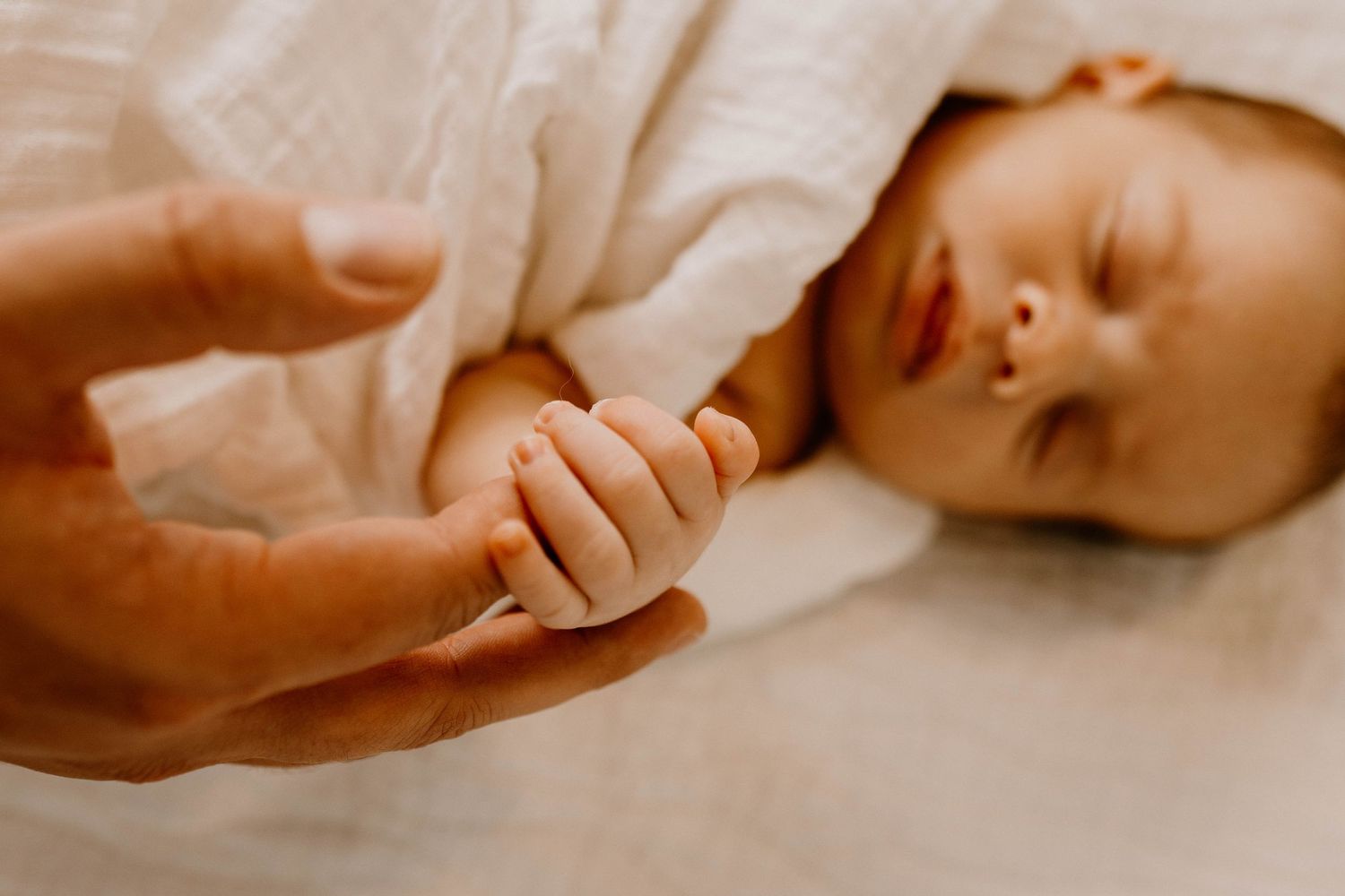Small sleeping baby holding an adult's hand while lying on white bedding in warm lighting.