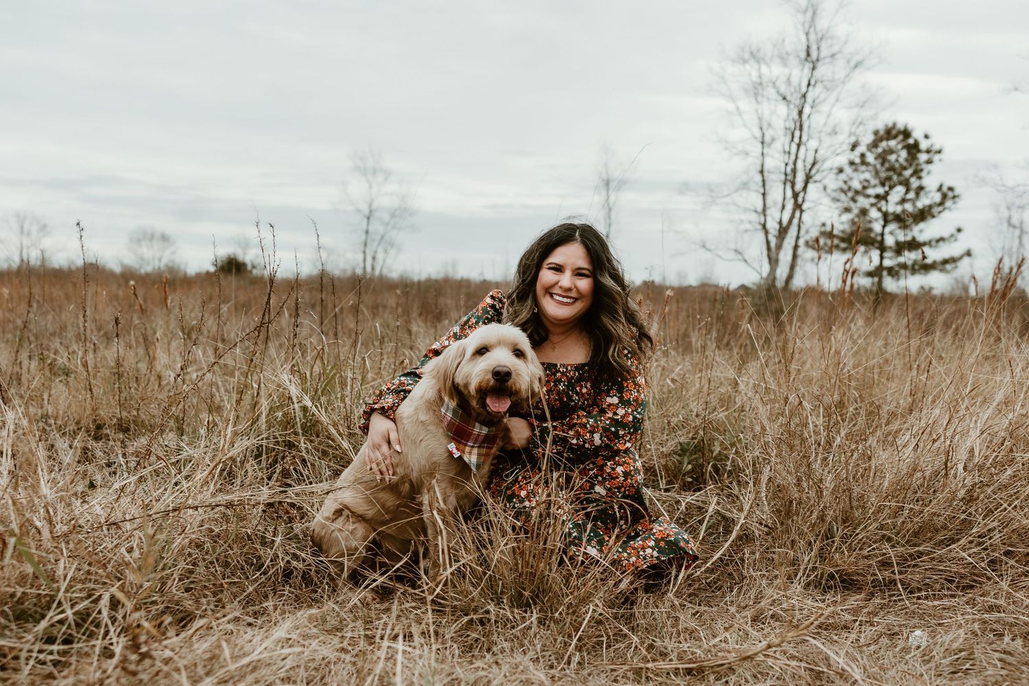 Person and goldendoodle dog sitting together in a tall grass field on an overcast autumn day.