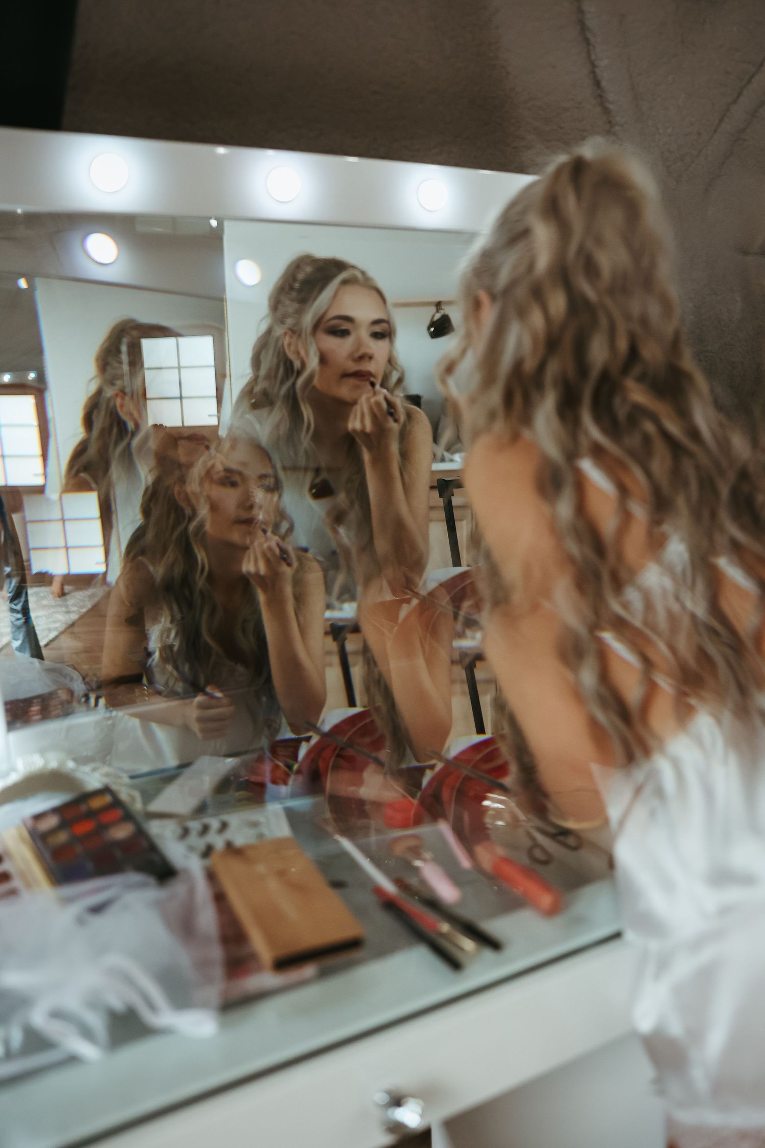 Person applies makeup while looking in a mirror surrounded by beauty products and cosmetics in a dressing room.