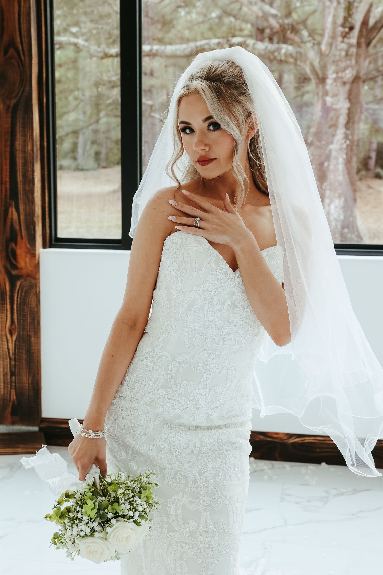 Stunning strapless lace wedding gown with flowing veil features delicate white floral bouquet and natural window lighting.