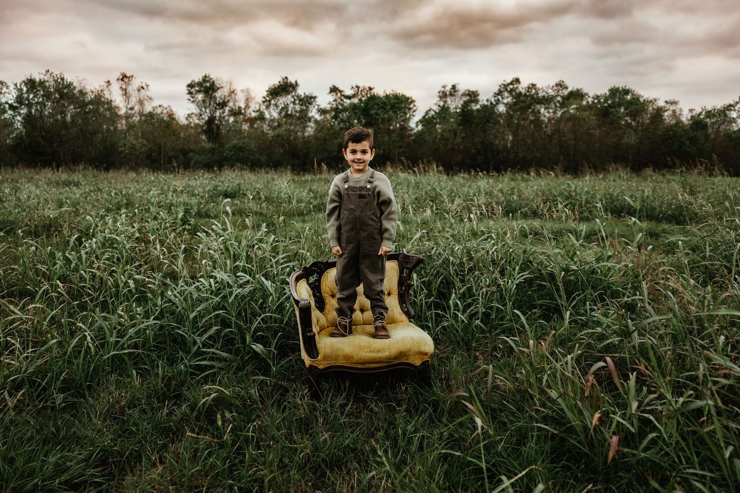A child stands on a yellow lawn mower in a lush green field with trees in the background under cloudy skies.
