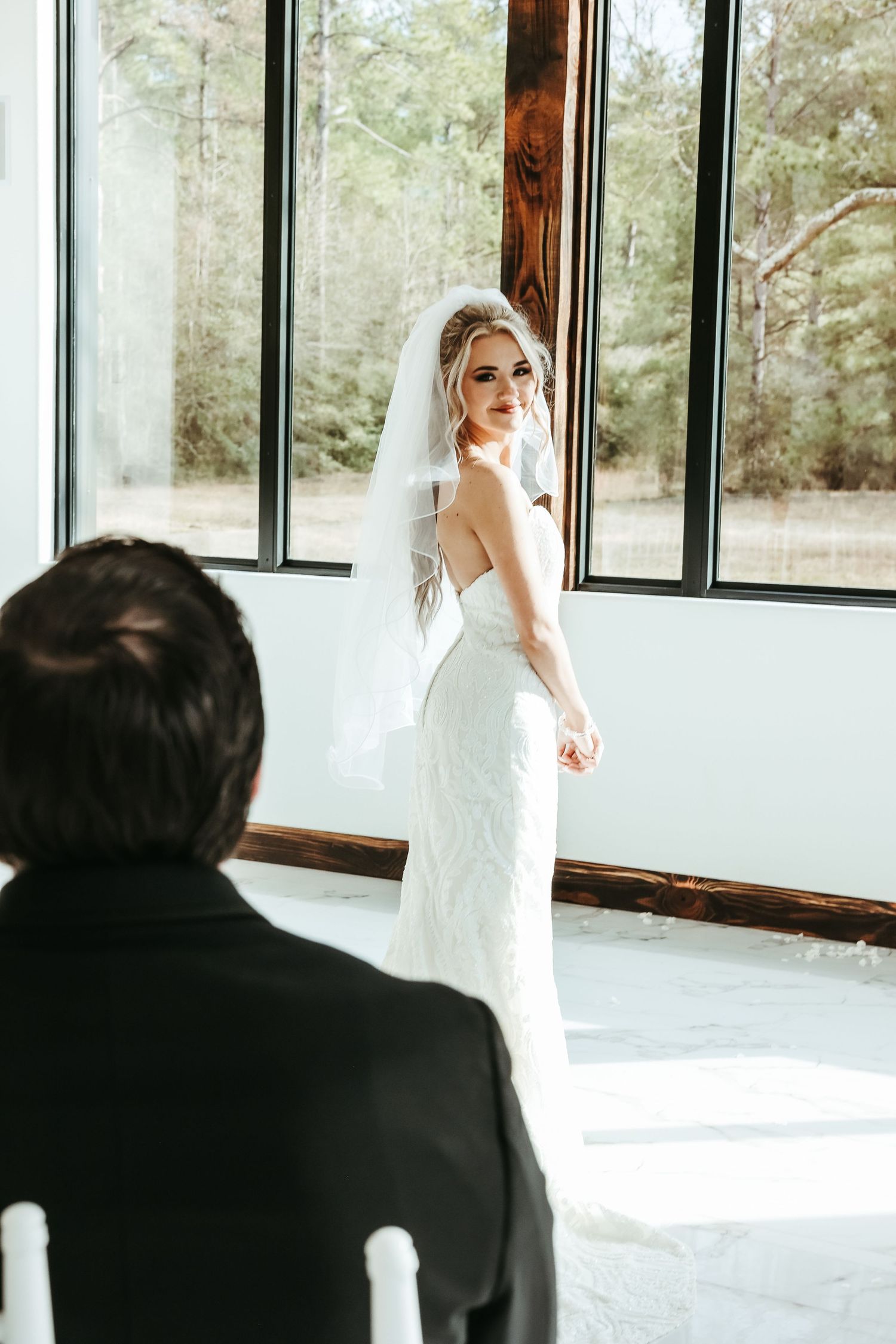 A bride in a fitted lace gown poses by sunlit windows during a first look moment at her wedding ceremony.