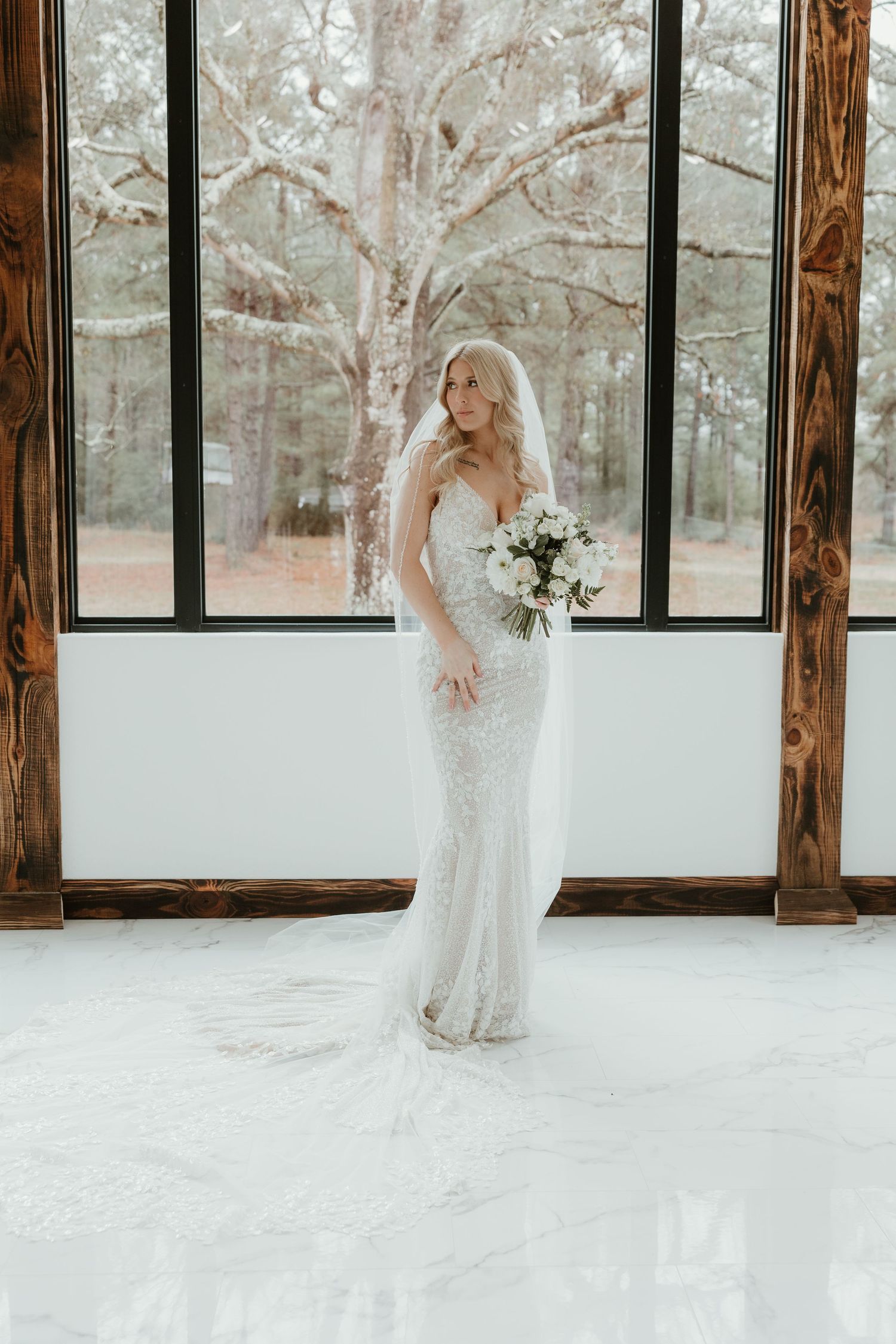 A bride in a fitted lace wedding dress poses by a large window in a rustic venue with snow-covered ground outside.