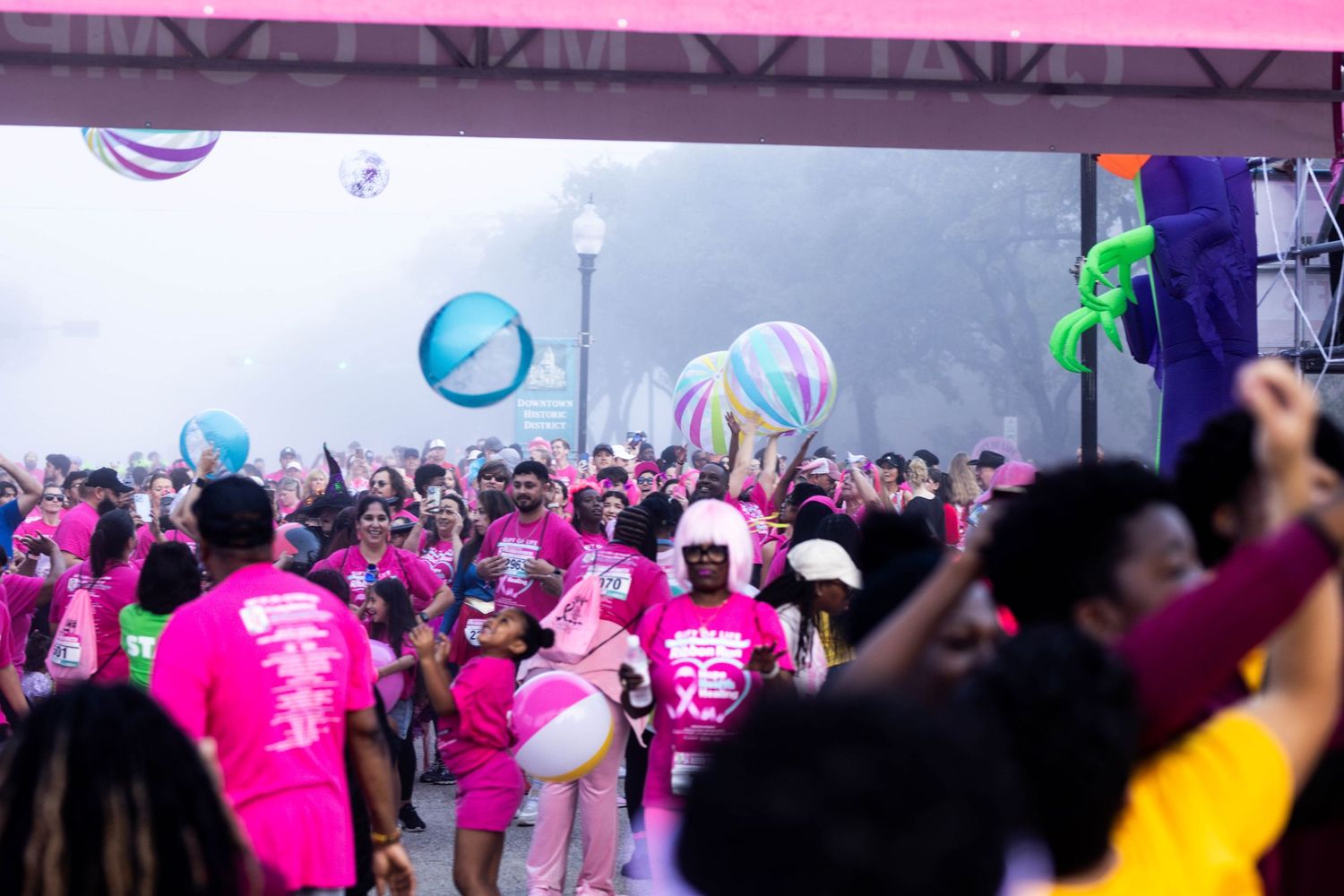 Large crowd wearing pink shirts throws colorful beach balls in the air during an outdoor event.