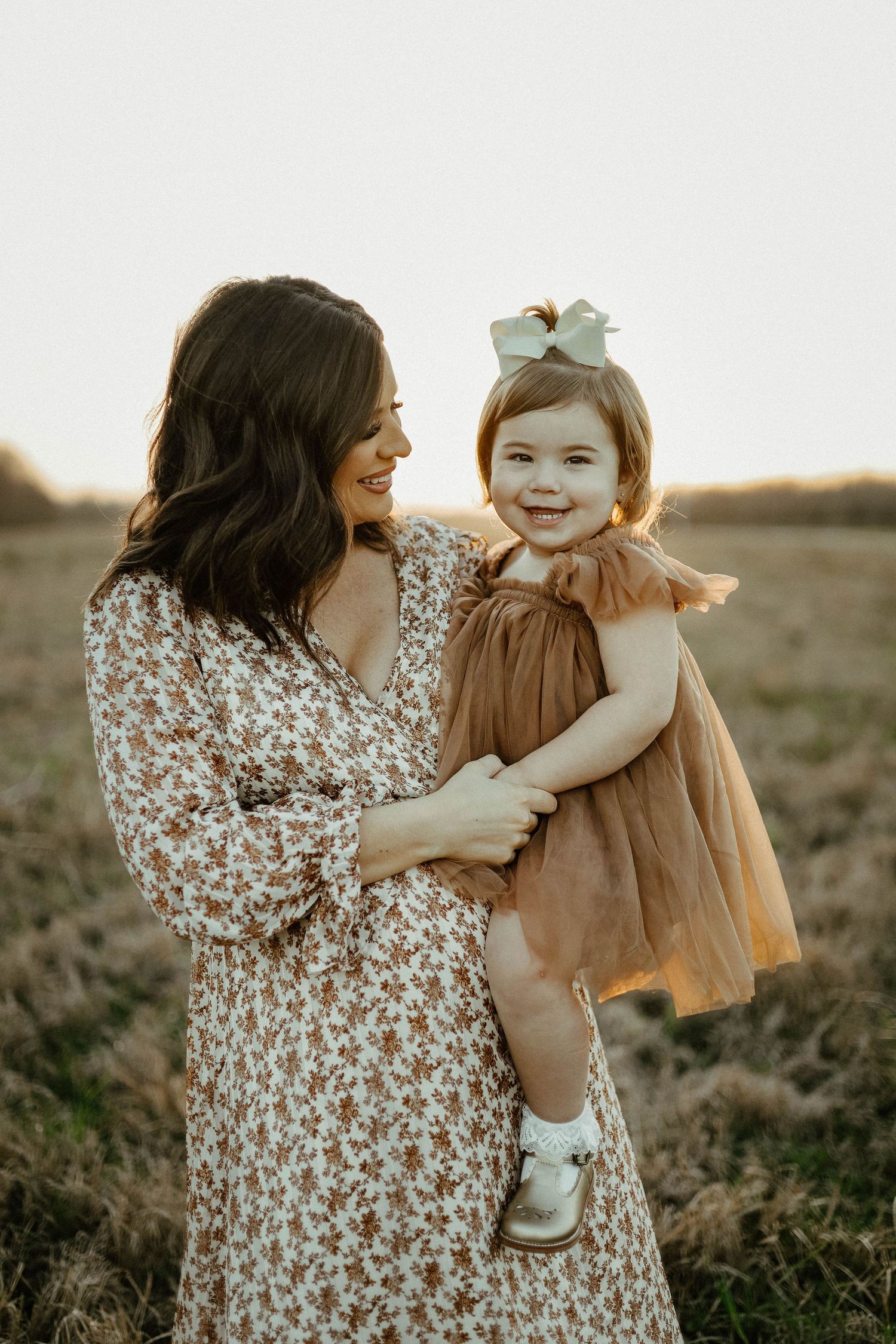Beautiful sunset portrait of mother and daughter in floral dress and brown dress standing in a golden field.