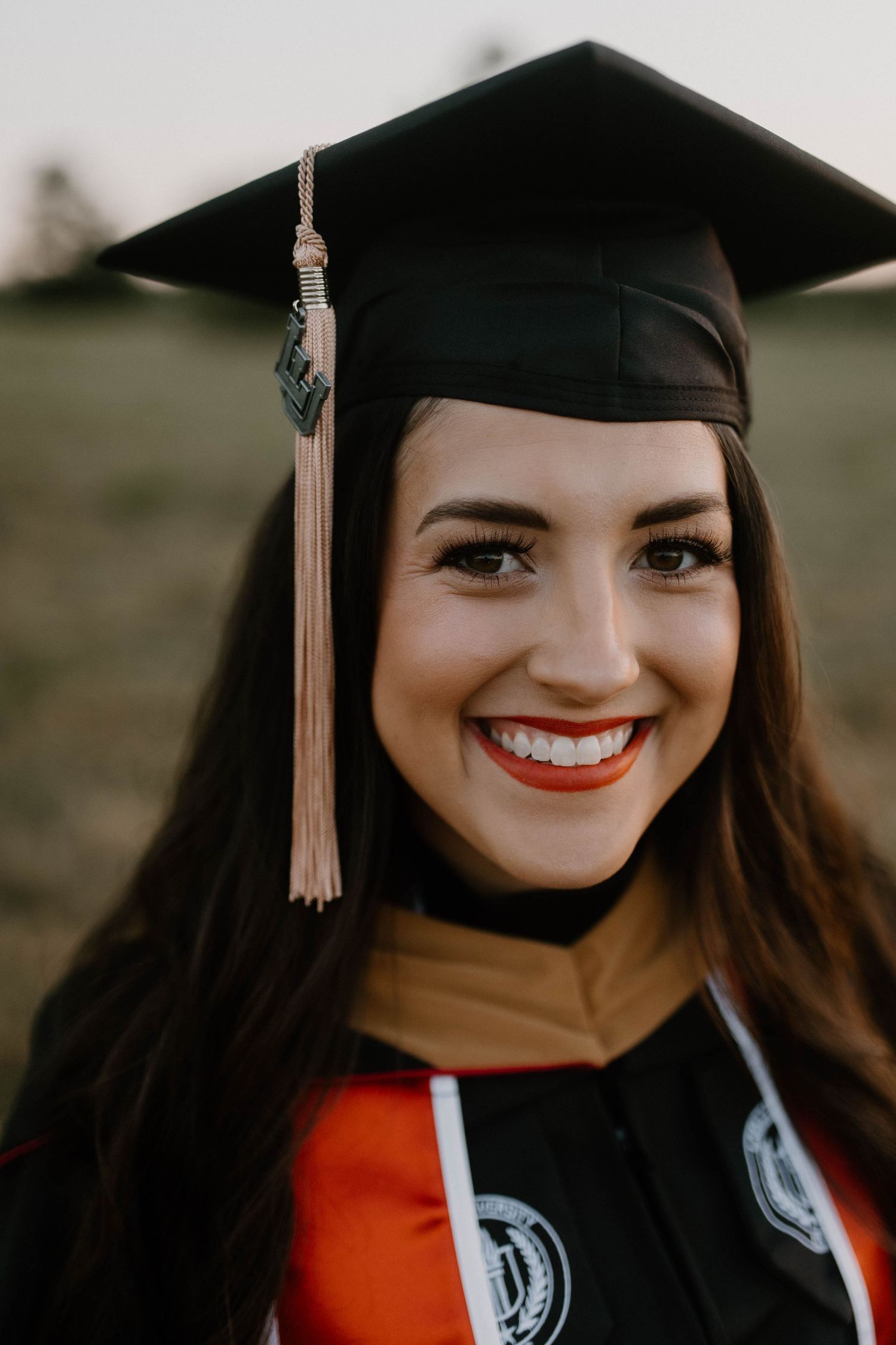Graduate student in cap and gown with red graduation stole smiles brightly in close-up graduation portrait.