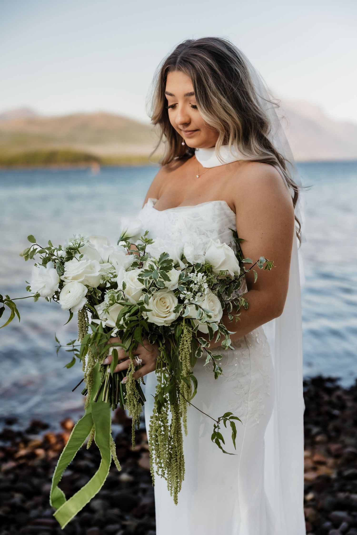A bride in a white dress holds a lush bouquet of white roses and cascading greenery against an ocean backdrop.