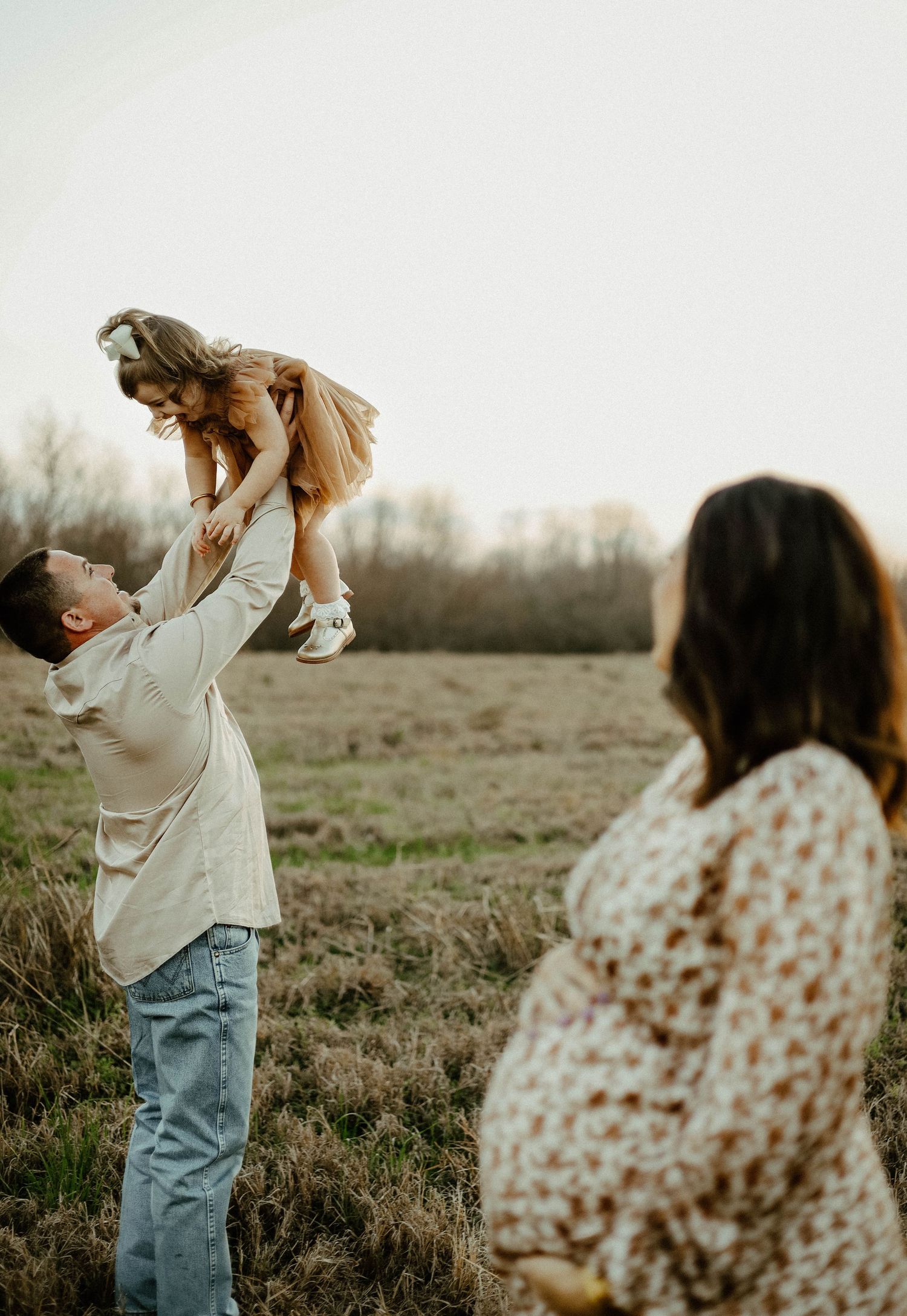 A person lifts a small child in the air in a field at dusk while another watches from the side.