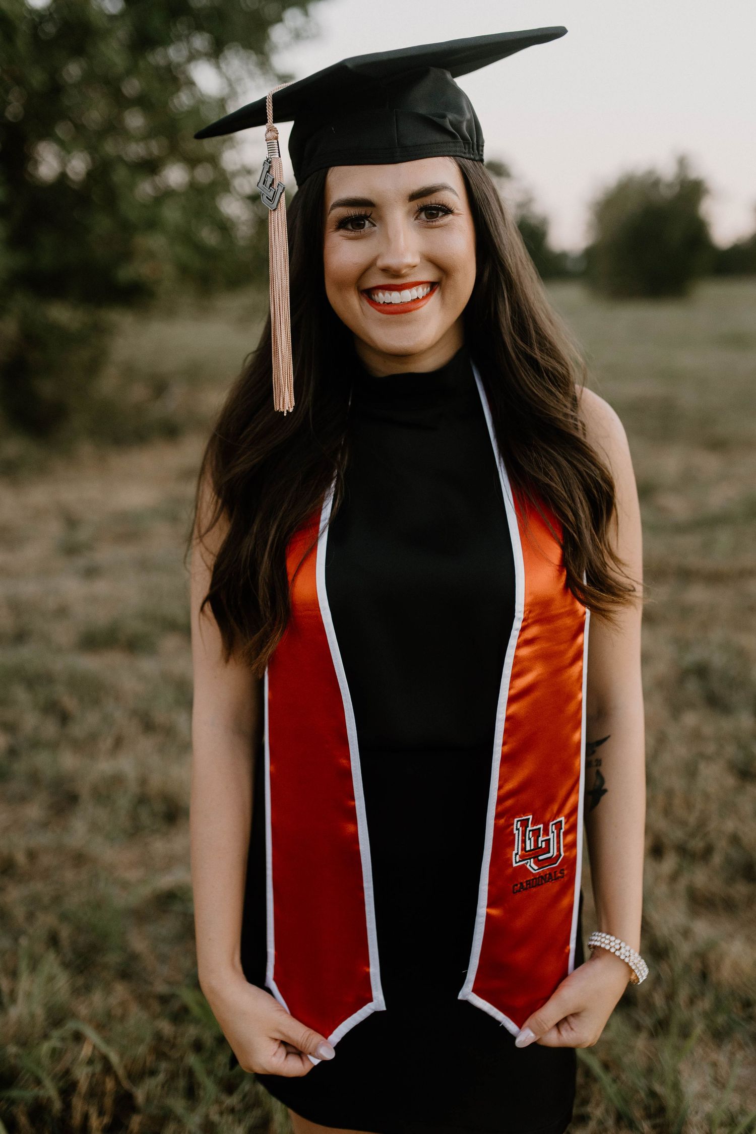 A graduate wearing a black cap and gown with red and white graduation stole stands in an outdoor field at sunset.