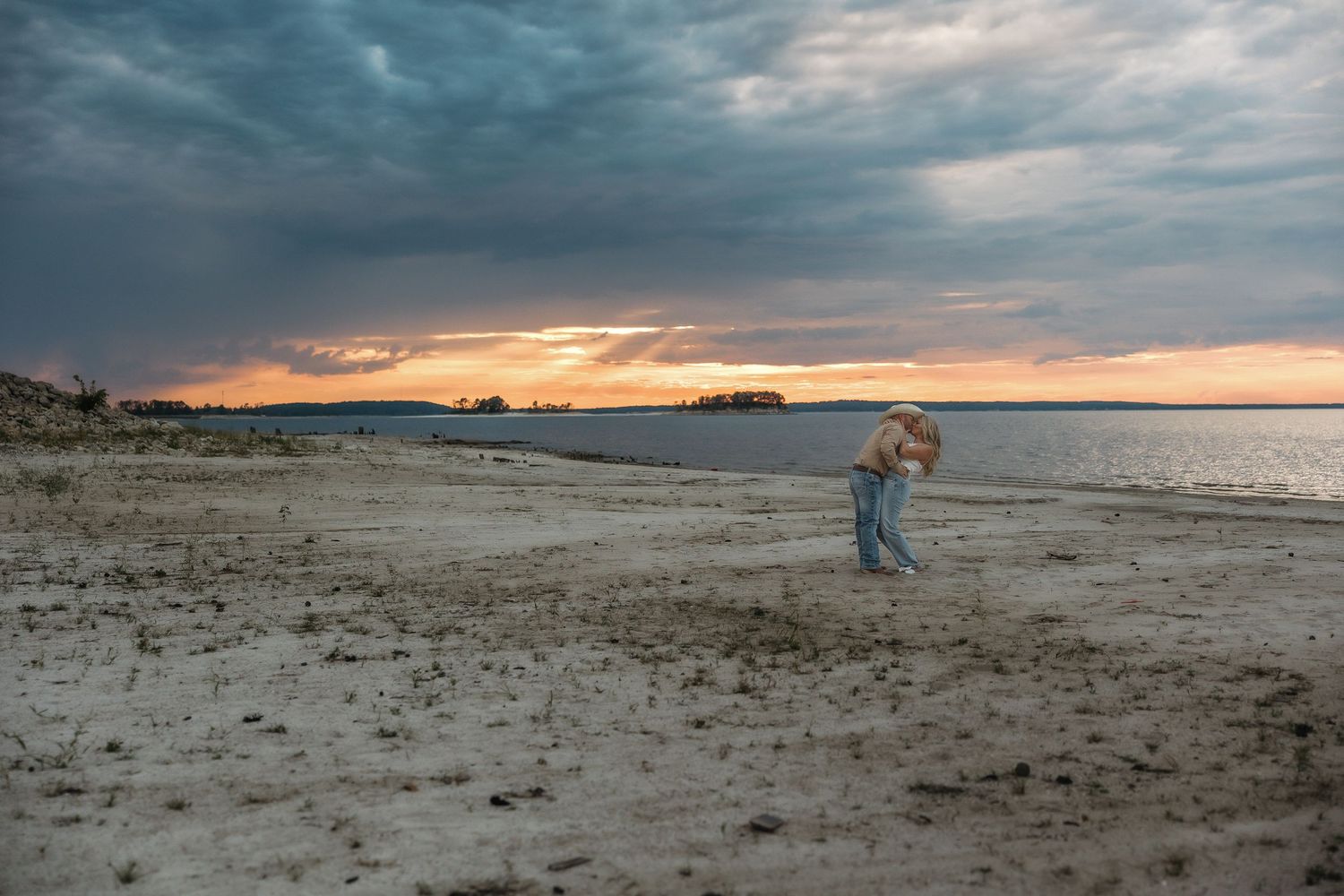 A couple walks hand in hand along a sandy beach at sunset with dramatic cloudy skies overhead.