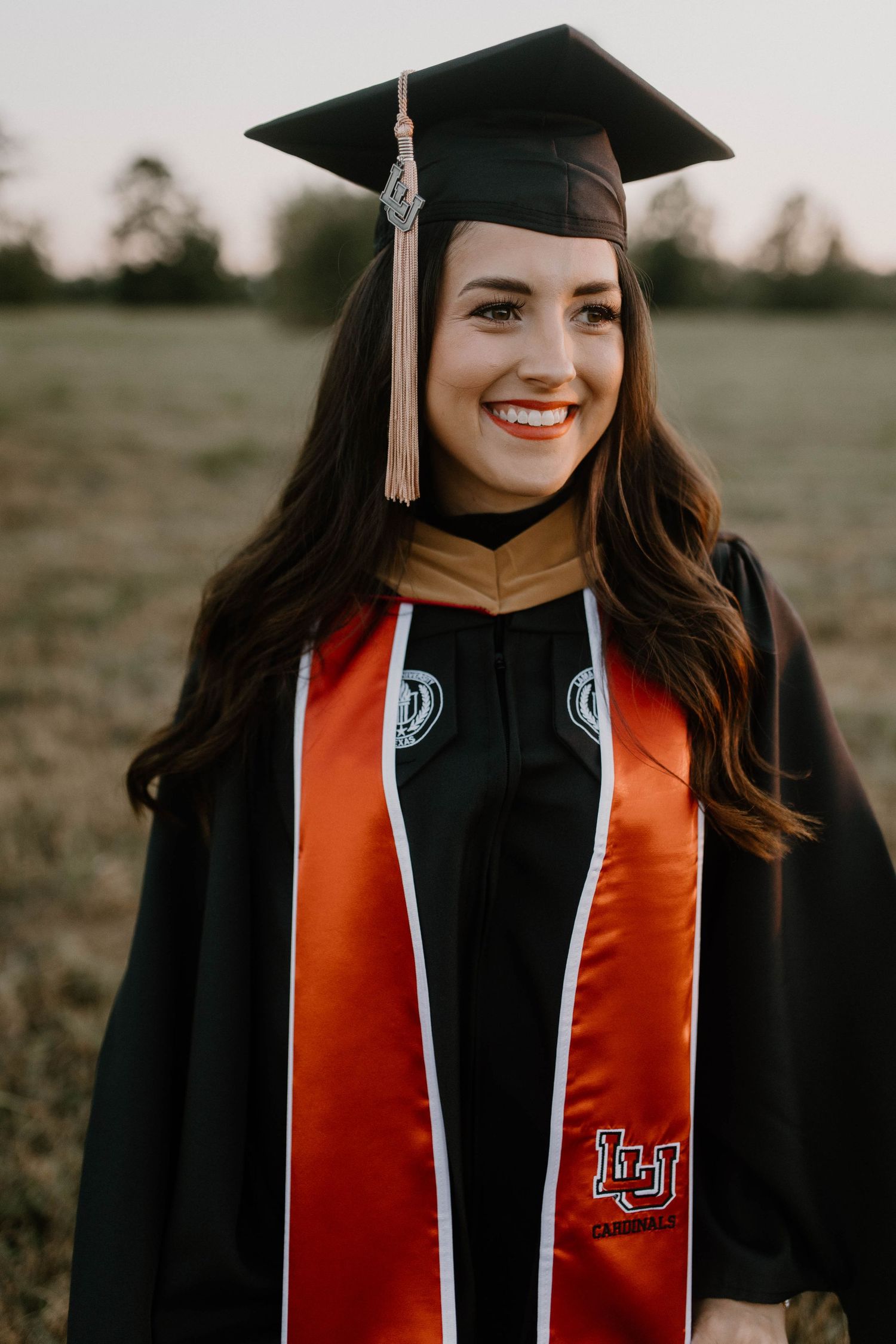 Graduate wearing black and orange academic regalia and graduation cap smiles during outdoor portrait session.