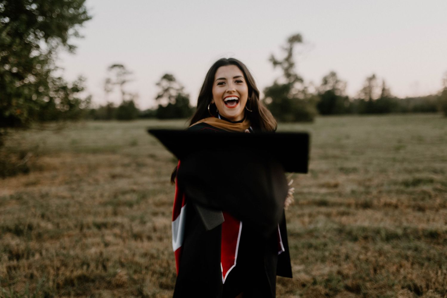 Graduate holding up diploma with joyful expression in outdoor field at sunset.