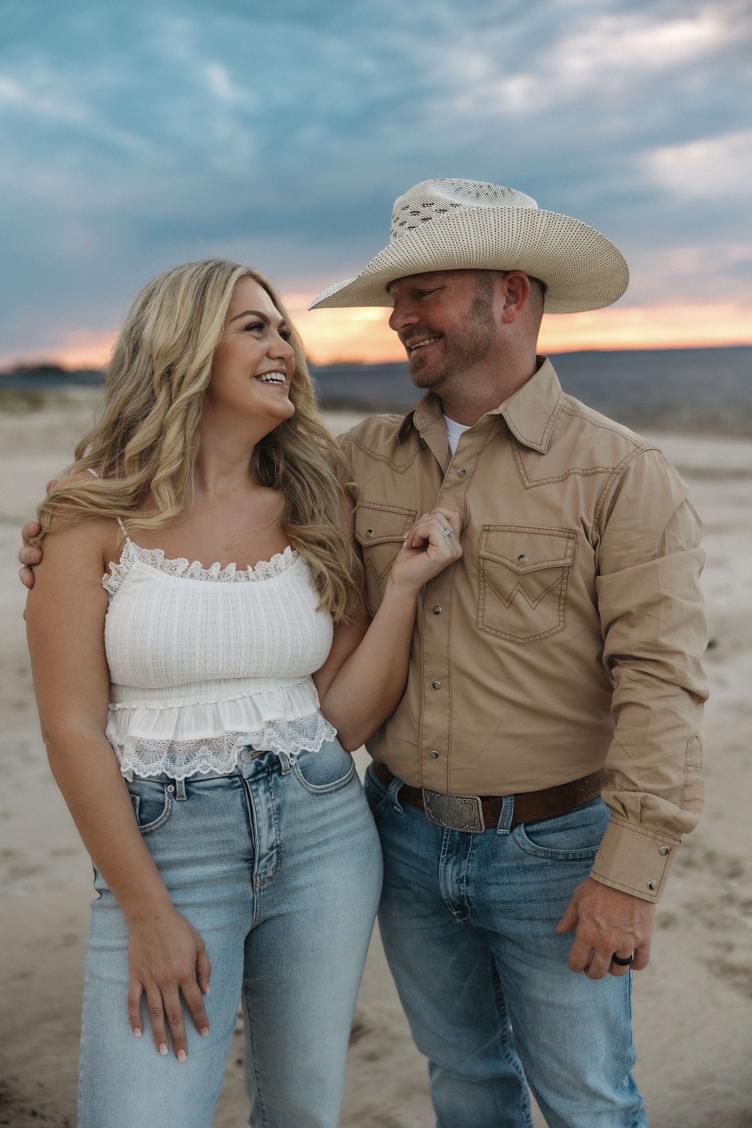 A romantic couple in western attire laughs together against a sunset desert backdrop.