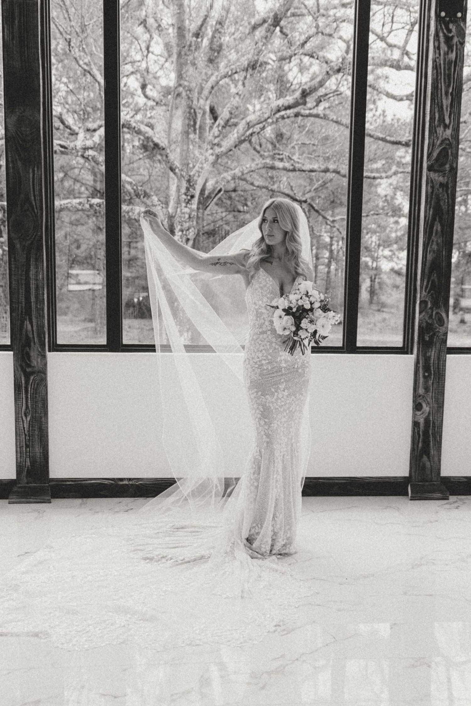 A bride in a fitted lace gown holds her flowing veil by a window in a black and white bridal portrait.