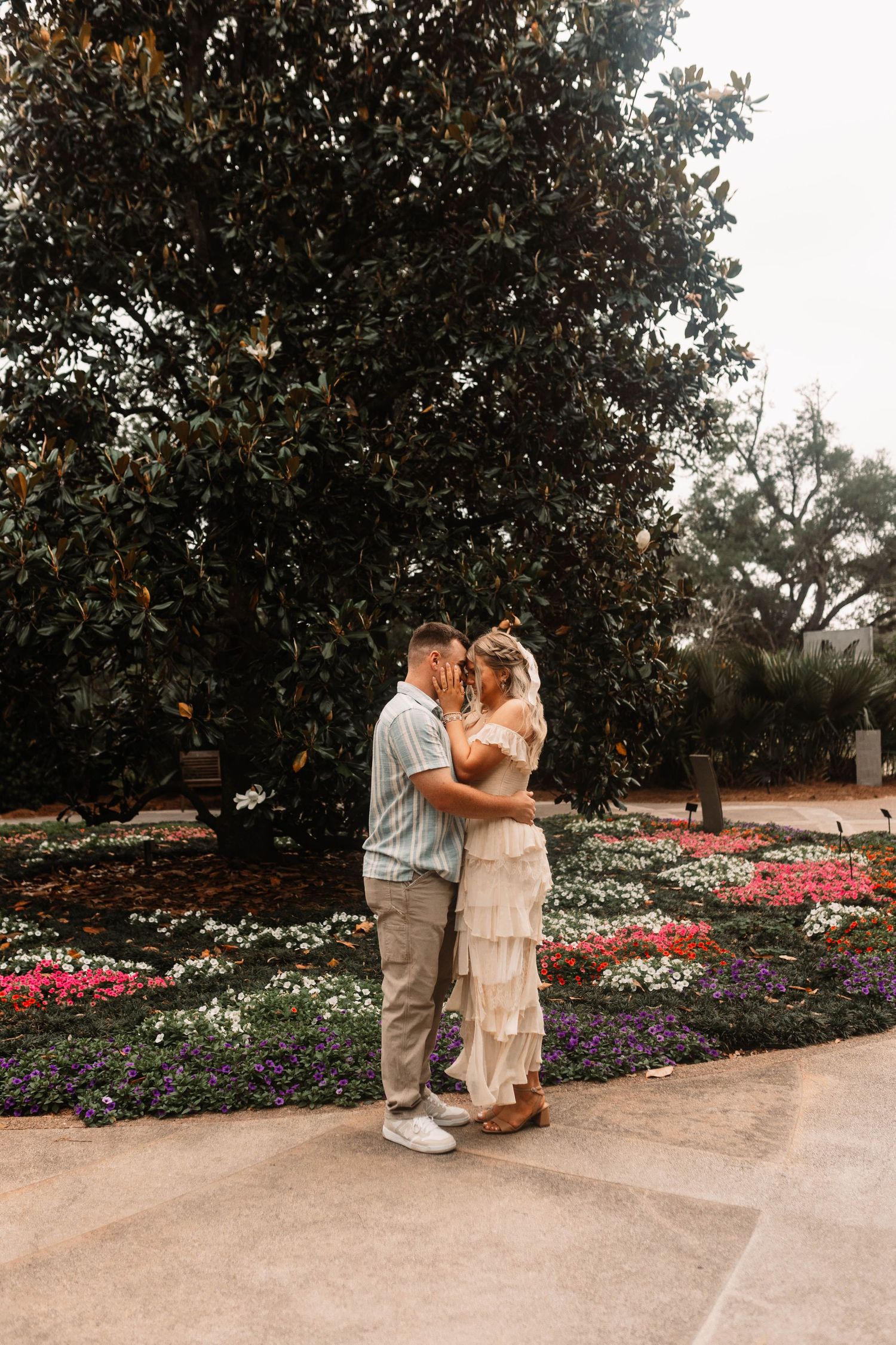 A couple shares a romantic moment in a garden with colorful flower beds and magnolia trees.