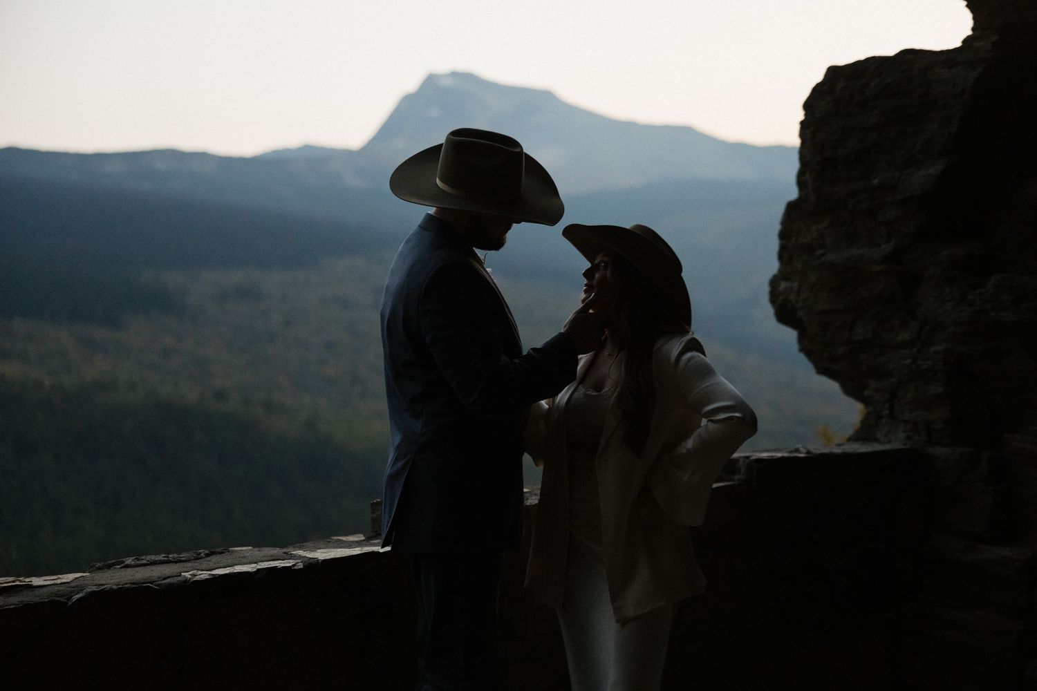 Silhouetted figures stand against a backdrop of majestic mountain ranges during sunset.