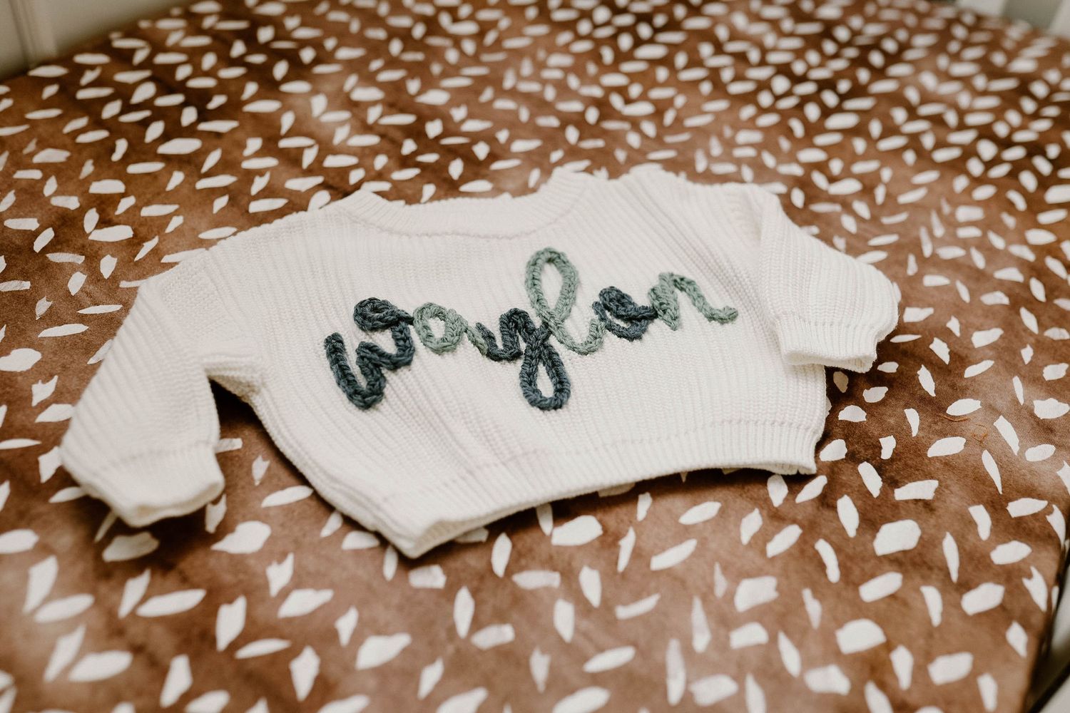 Small white baby sweater with green lettering embroidered on it lies on a patterned brown and white fabric.