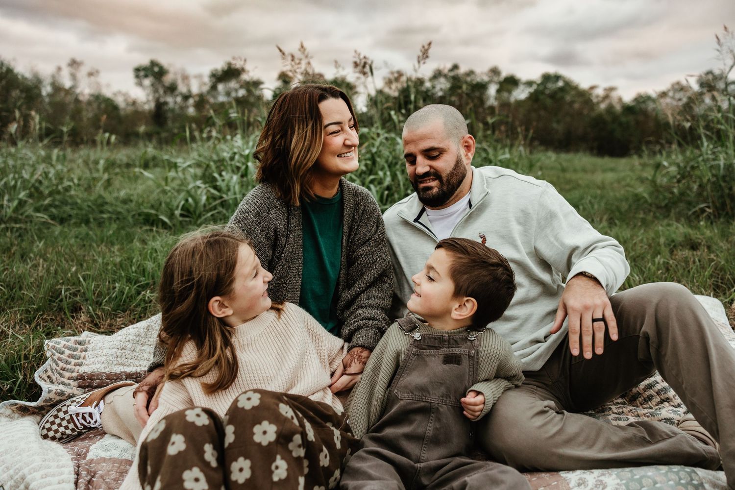 A happy family shares warm laughs while relaxing together on a blanket in a green natural setting at sunset.