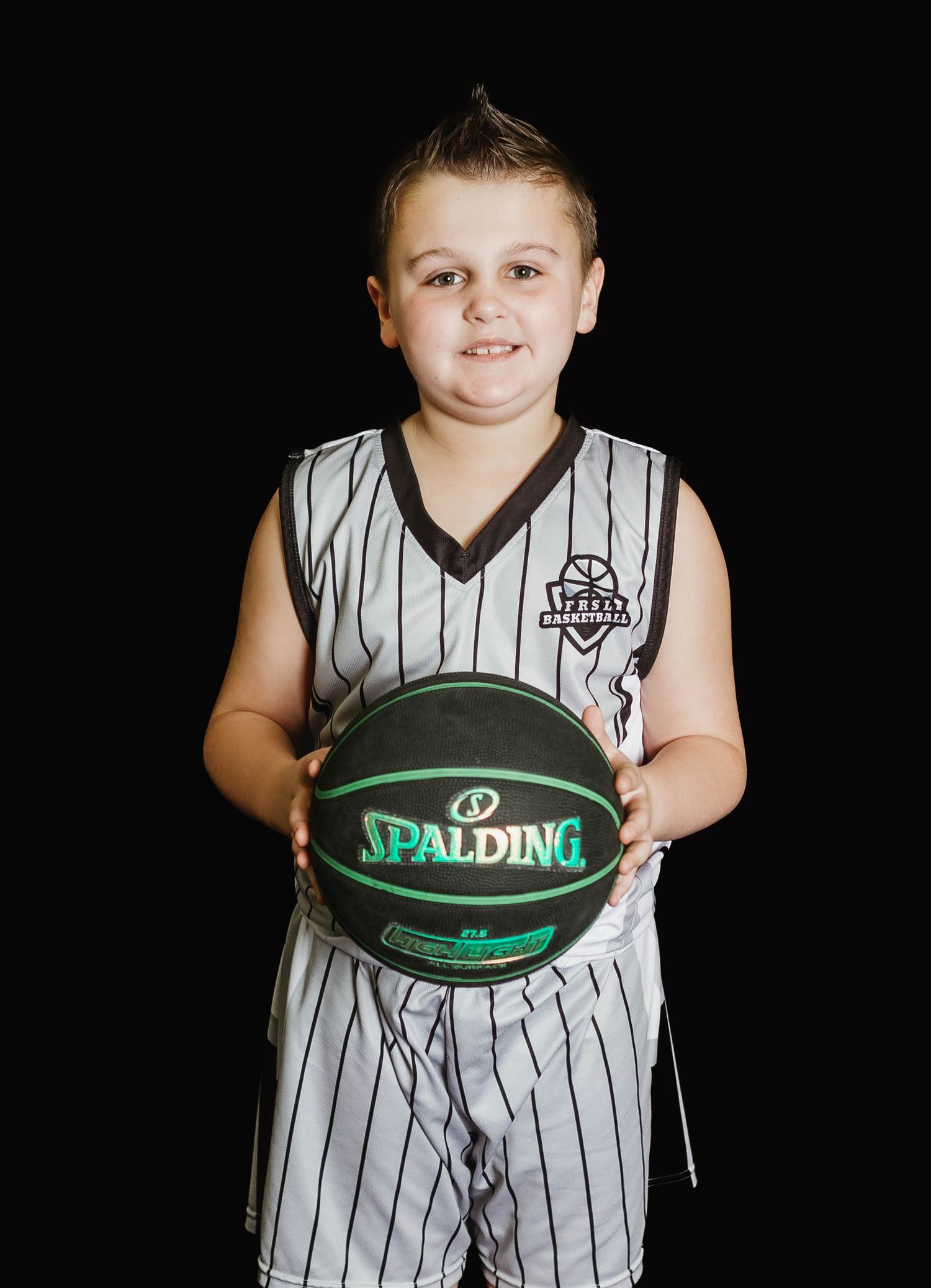 Young basketball player in a striped jersey holding a green Spalding basketball against dark background.